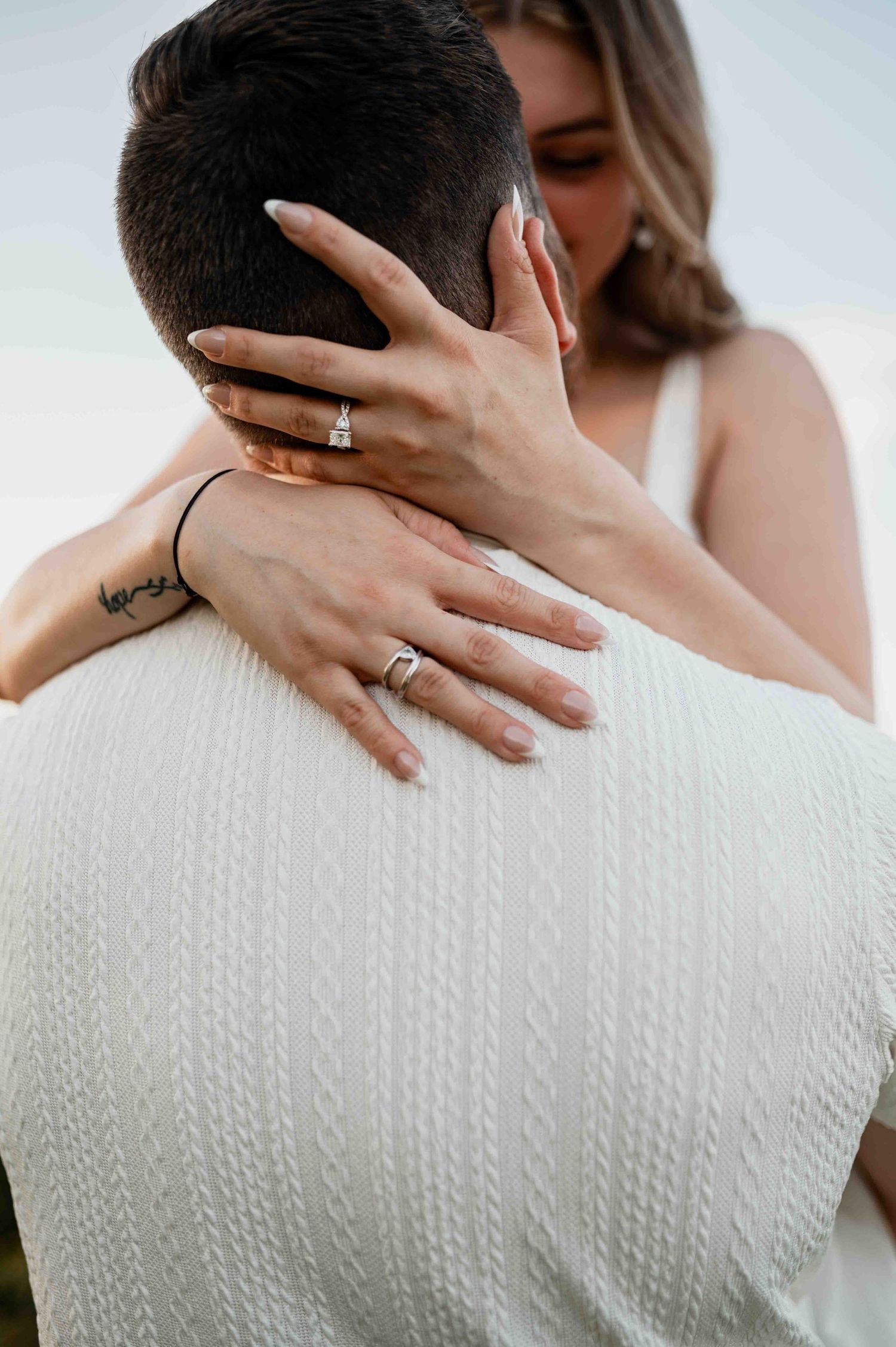 Couple embracing in white outfits, showcasing elegant engagement rings on the bride's hand during a romantic moment.