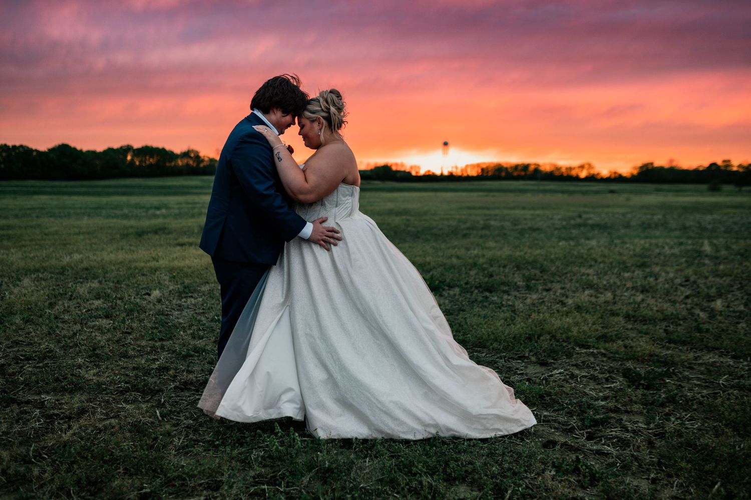 Bride and groom share a romantic kiss at sunset on a lush green field, with a stunning red and orange sky backdrop.