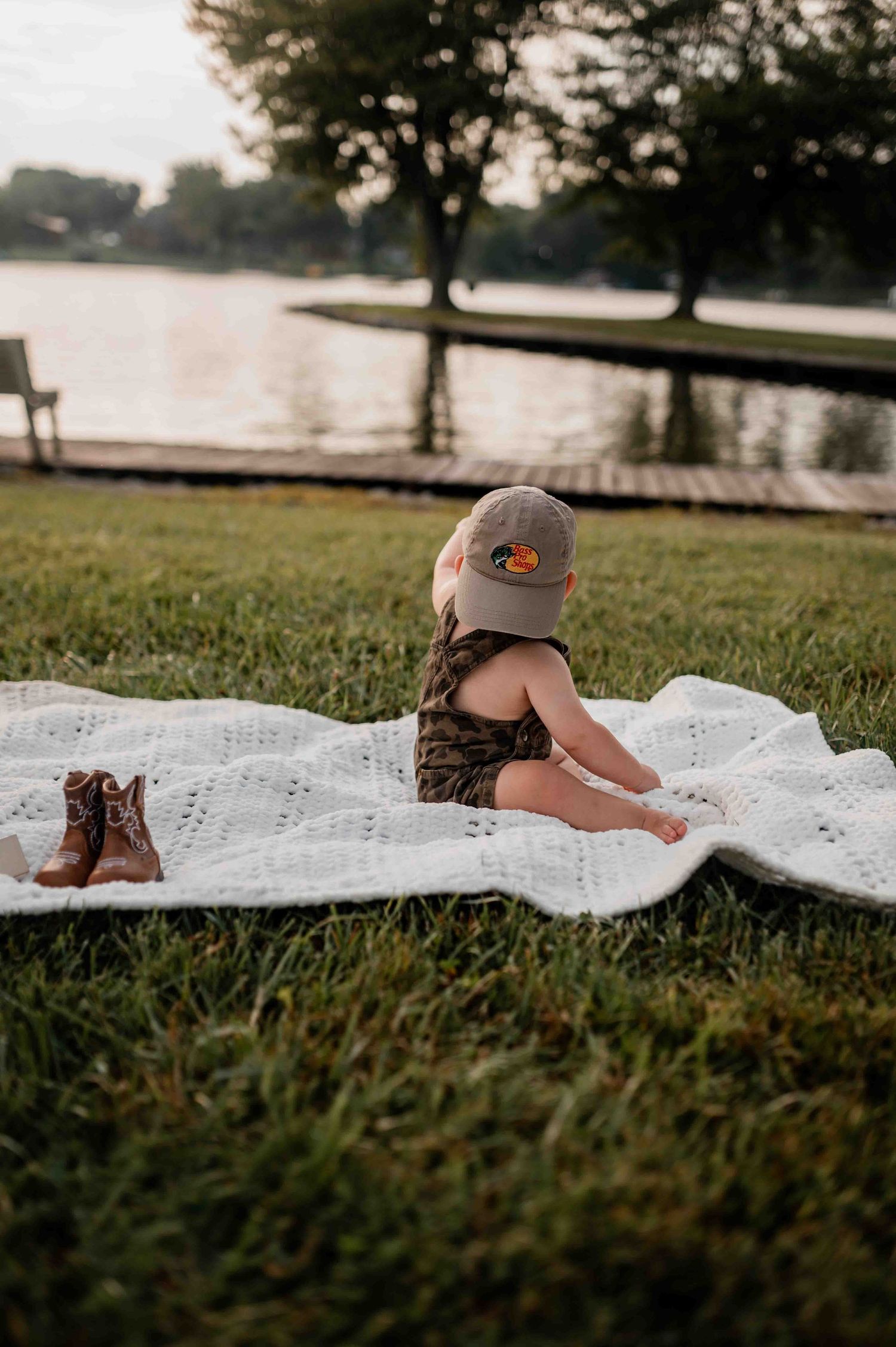 Low angle view of baby crawling on white blanket in park setting with lake in background.