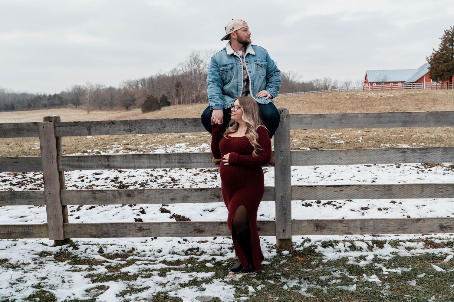 Couple poses at rustic farm fence in winter, woman in red dress with baby bump, man in denim jacket seated above.