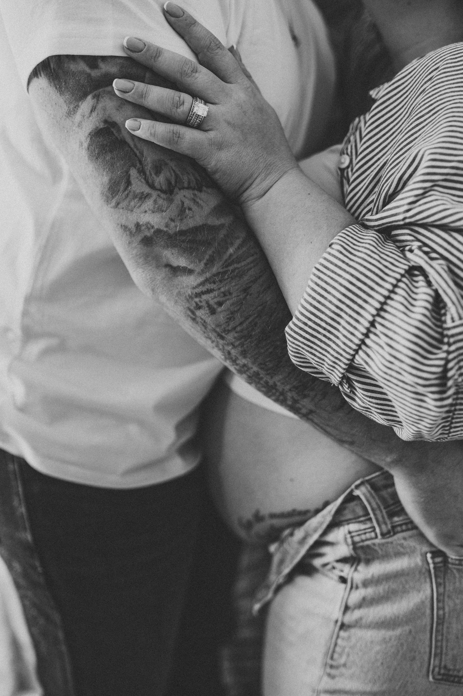 Black and white photo of a pregnant couple, man with tattooed arm and woman wearing a ring, embracing tenderly.