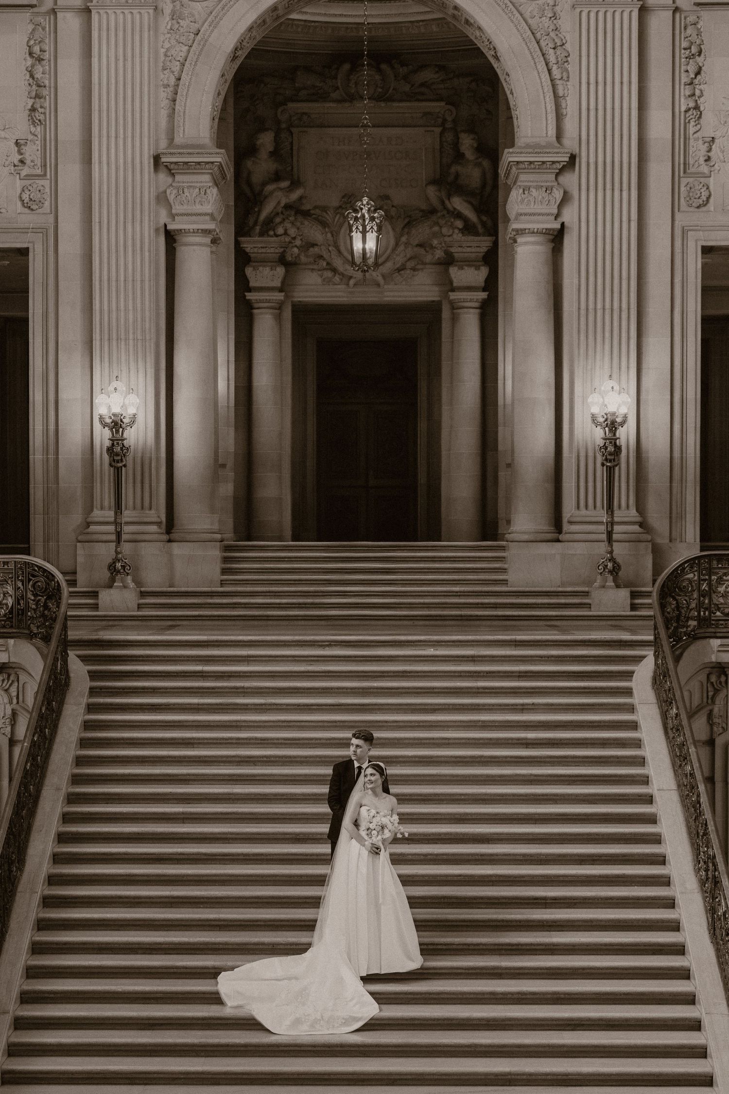 Bride and groom embrace on grand staircase of ornate wedding venue in elegant black and white photo.