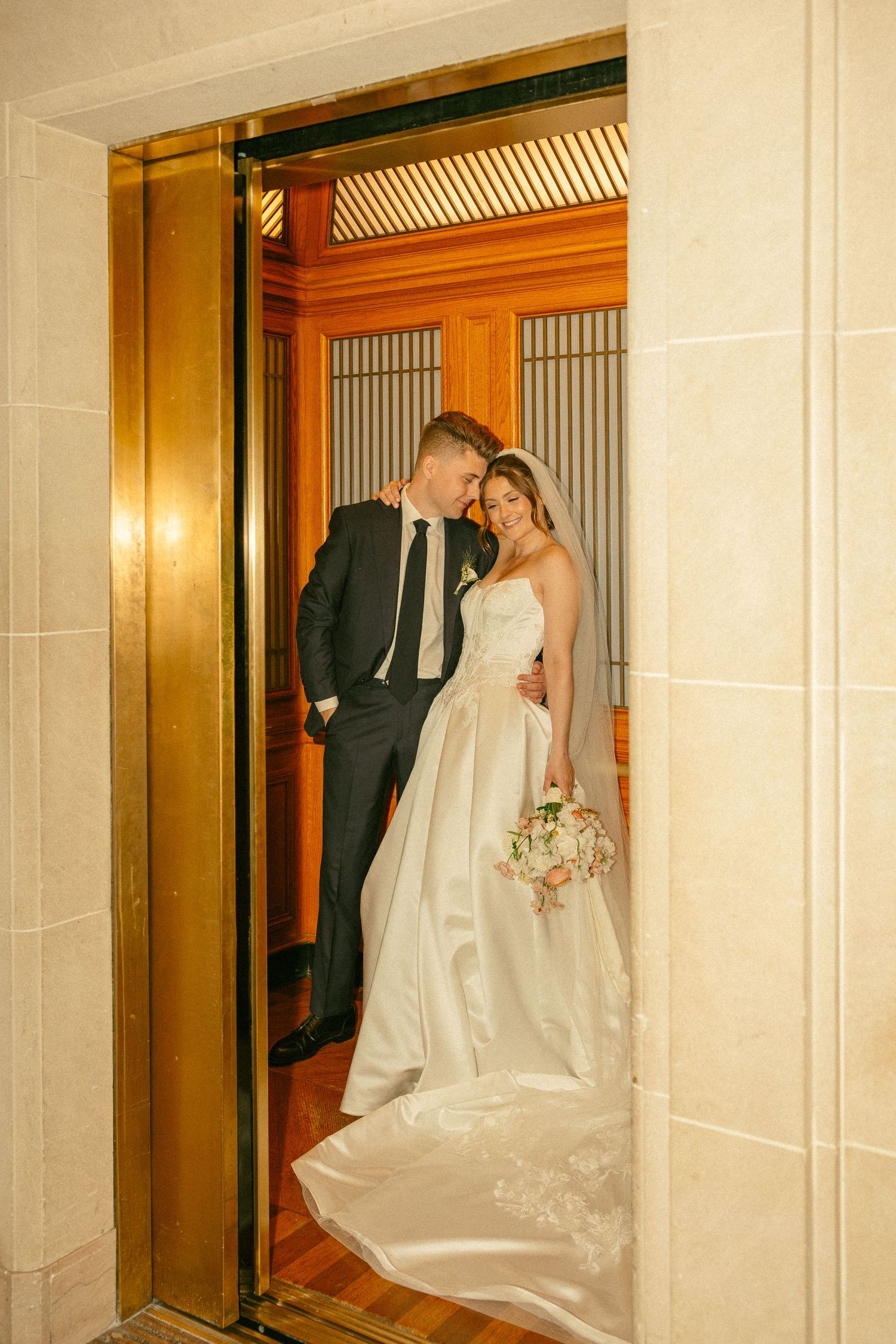 Bride and groom posing romantically in a gold elevator, bride in white ballgown holding bouquet, groom in dark suit.