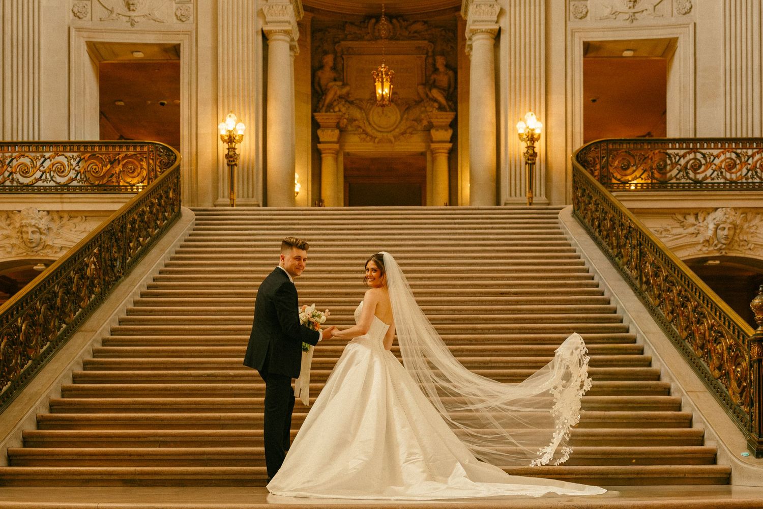 Bride and groom exchange vows on grand staircase of San Francisco City Hall in elegant wedding attire.
