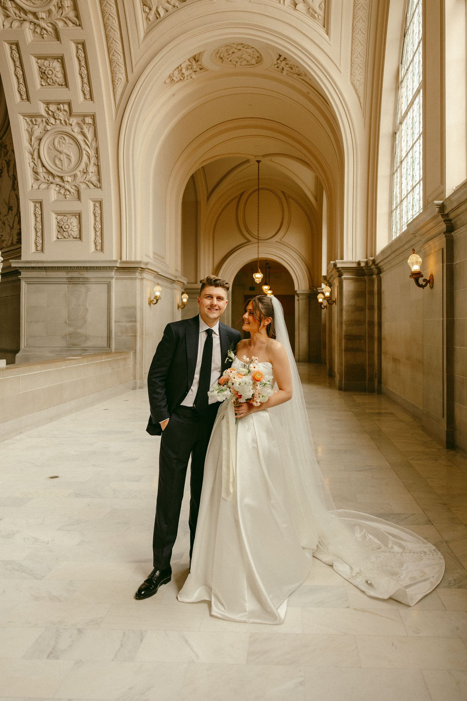 Bride and groom pose together in stunning arched marble corridor, bride holding bouquet in white gown.