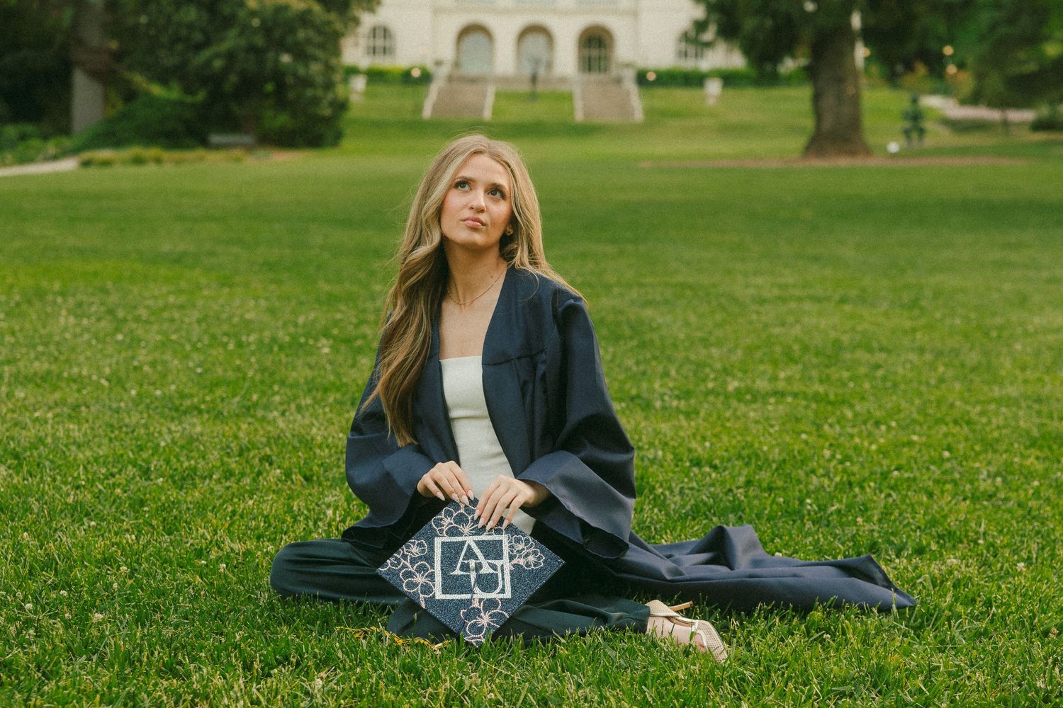 A blonde woman in graduation cap and gown sits on green grass holding her decorated mortarboard outdoors.