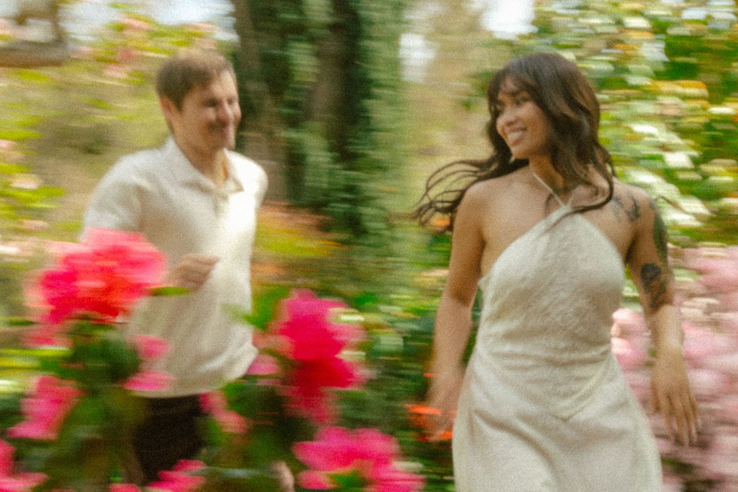 Couple in wedding attire running joyfully through a lush garden with pink flowers, motion blur effect.