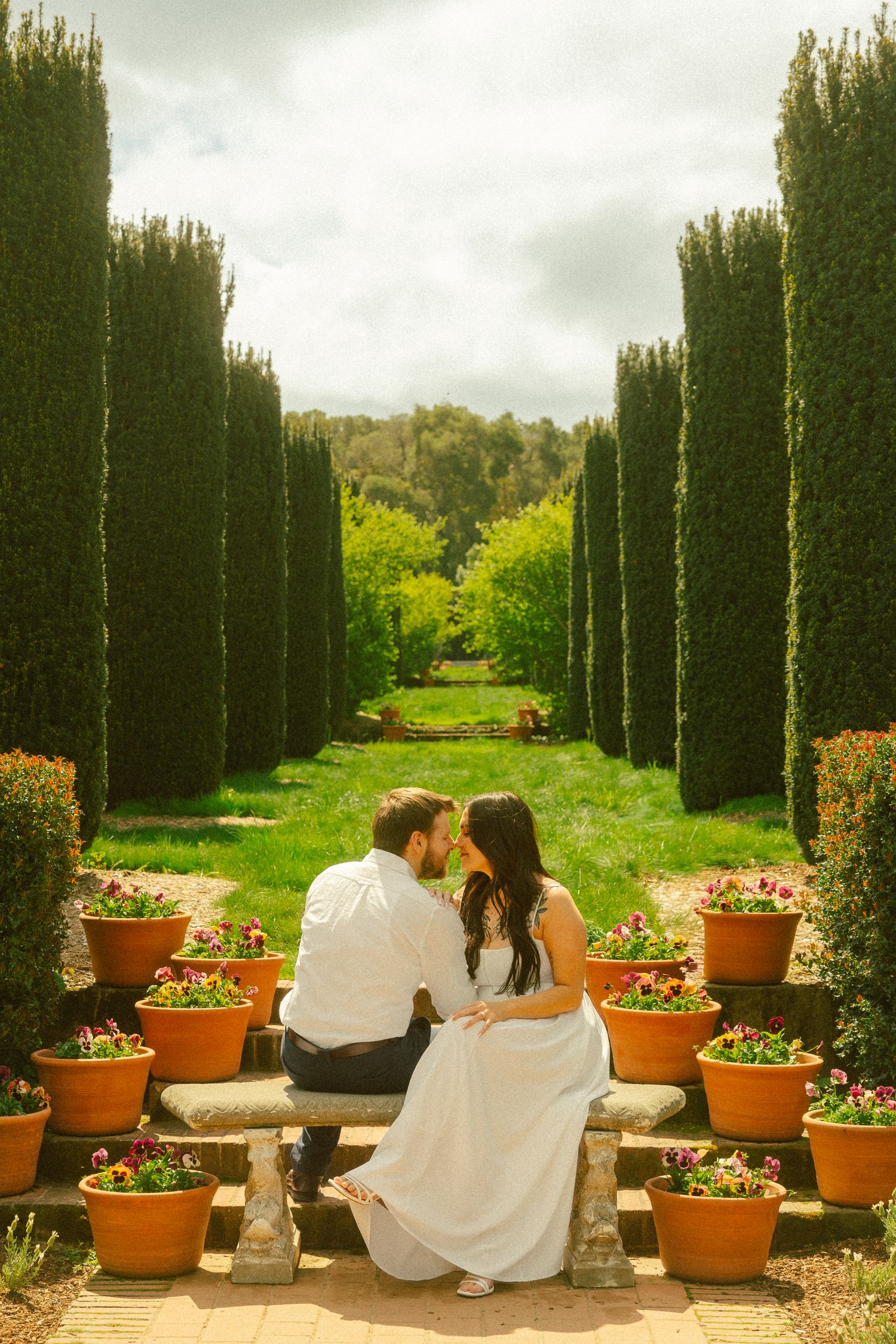 Couple sitting on stone bench in formal garden with tall cypress trees and terracotta flower pots.