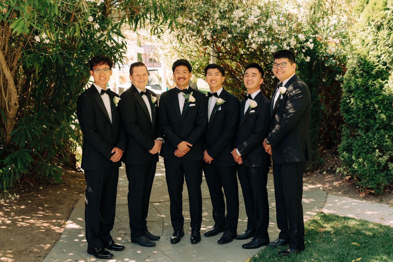 Six groomsmen in black tuxedos with bow ties and boutonnieres posing outdoors at a wedding.