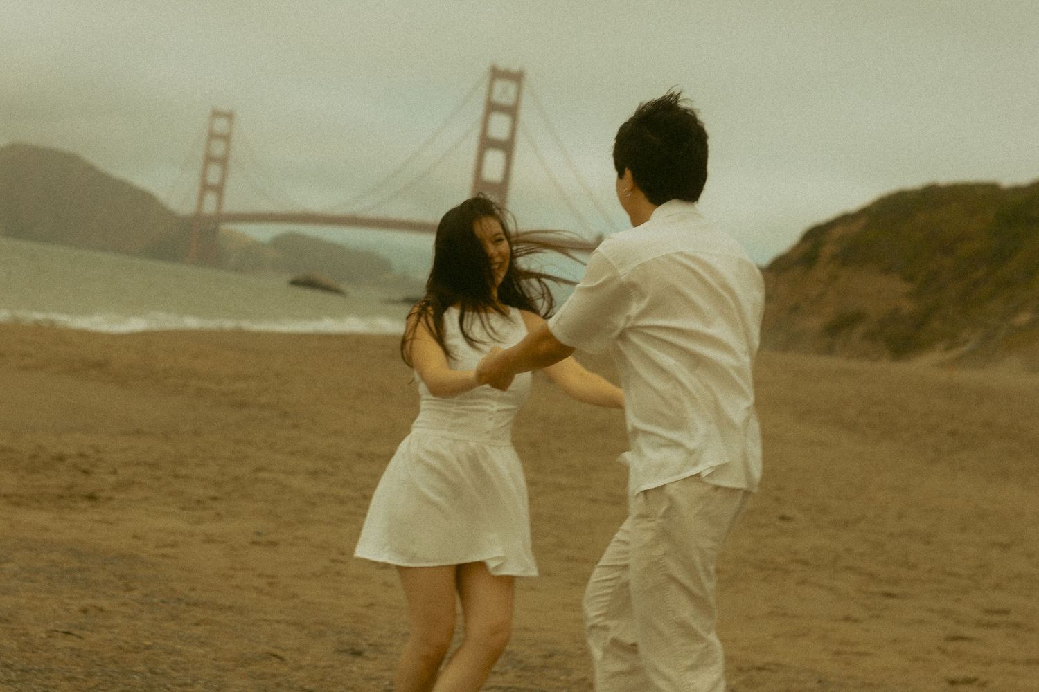 Couple dancing on Baker Beach with the Golden Gate Bridge in the background on a cloudy day.