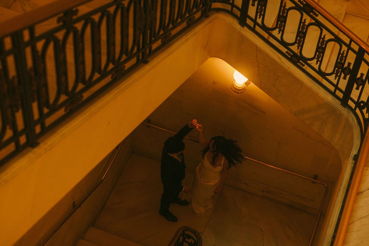 Aerial view of a couple dancing on ornate staircase under warm golden light at night.