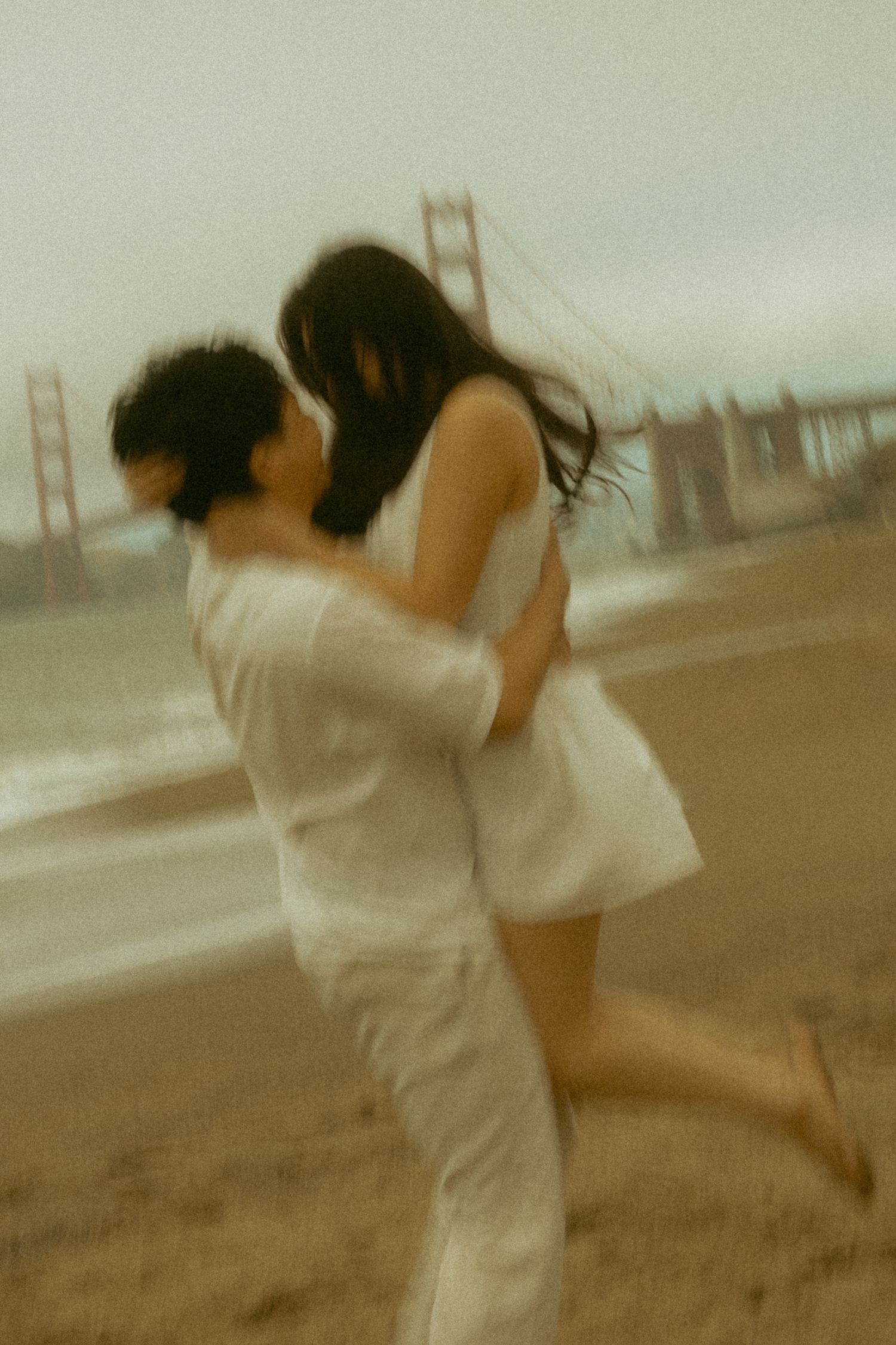 Couple embracing on Baker Beach with Golden Gate Bridge in background, captured in dreamy motion blur.