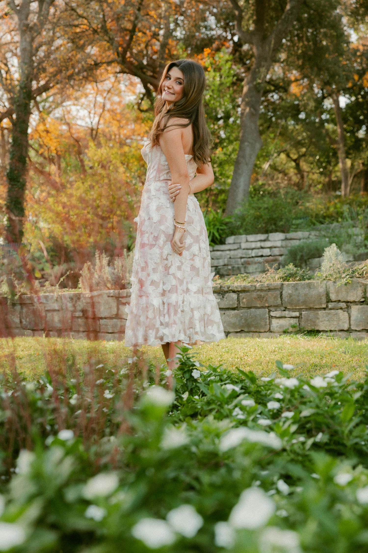 A floral dress flows gracefully against autumn foliage and stone walls in an outdoor garden setting.