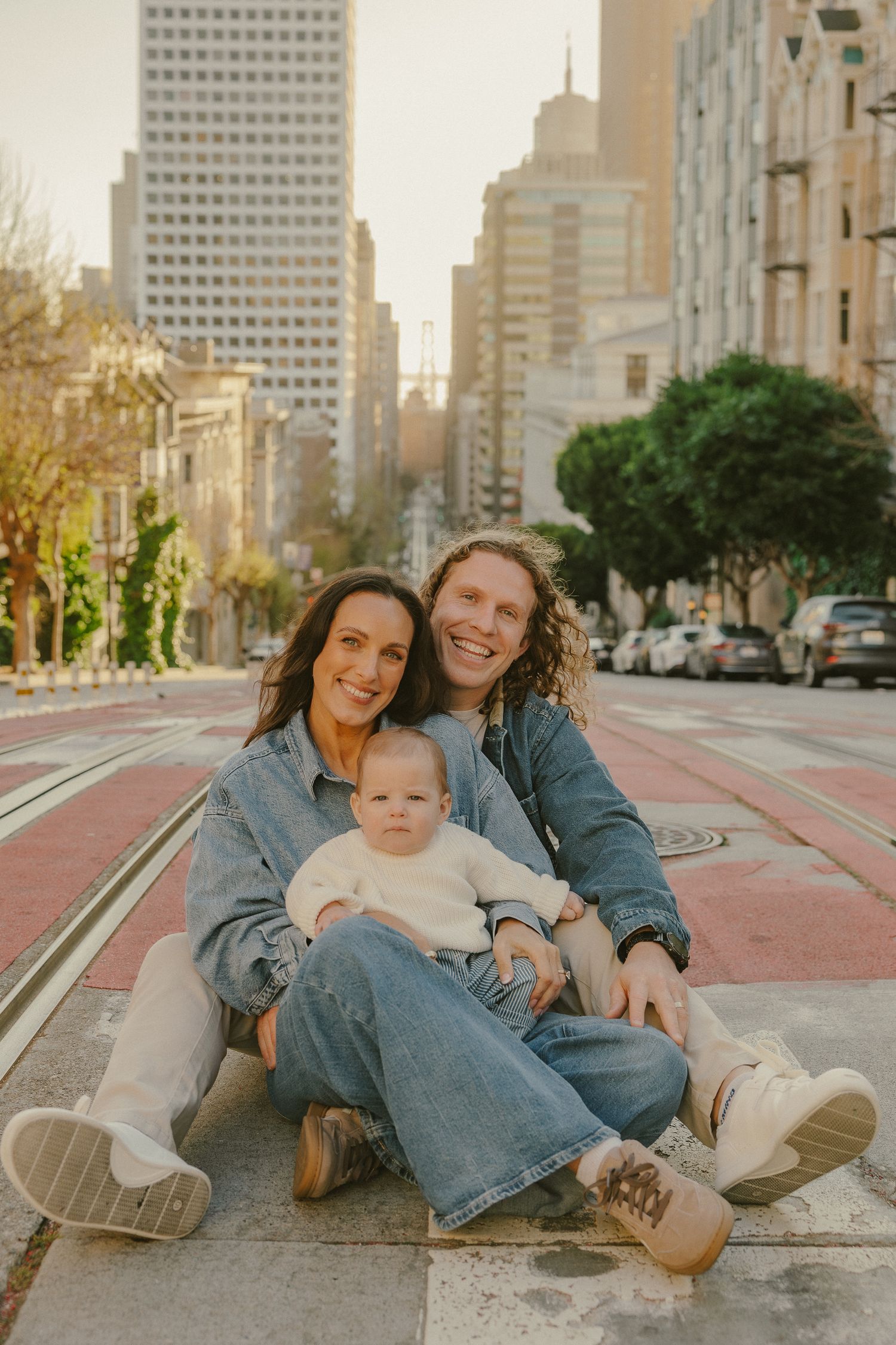 A happy family with a baby sits on a San Francisco street with city skyline in the background.