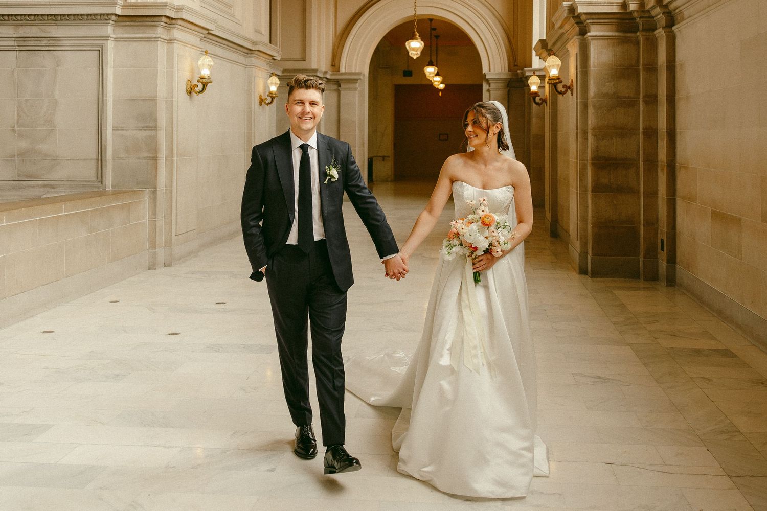 Bride and groom walk hand in hand through elegant marble hallway, bride holding colorful bouquet in white strapless gown.