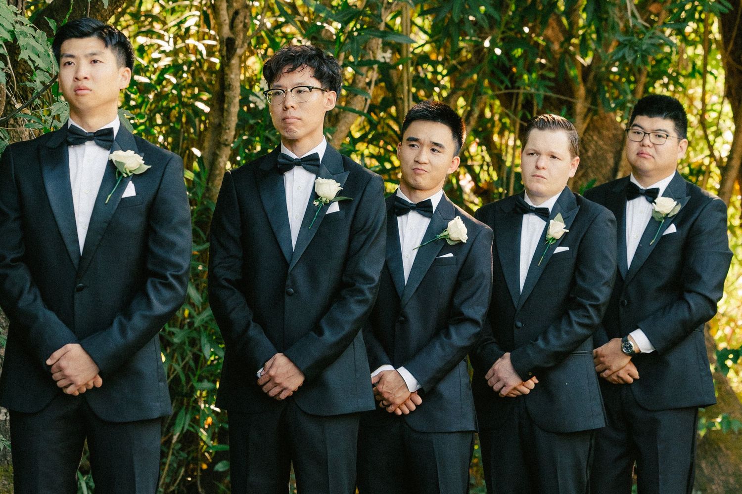 Five groomsmen in black tuxedos with white boutonnieres stand together outdoors at a wedding ceremony.
