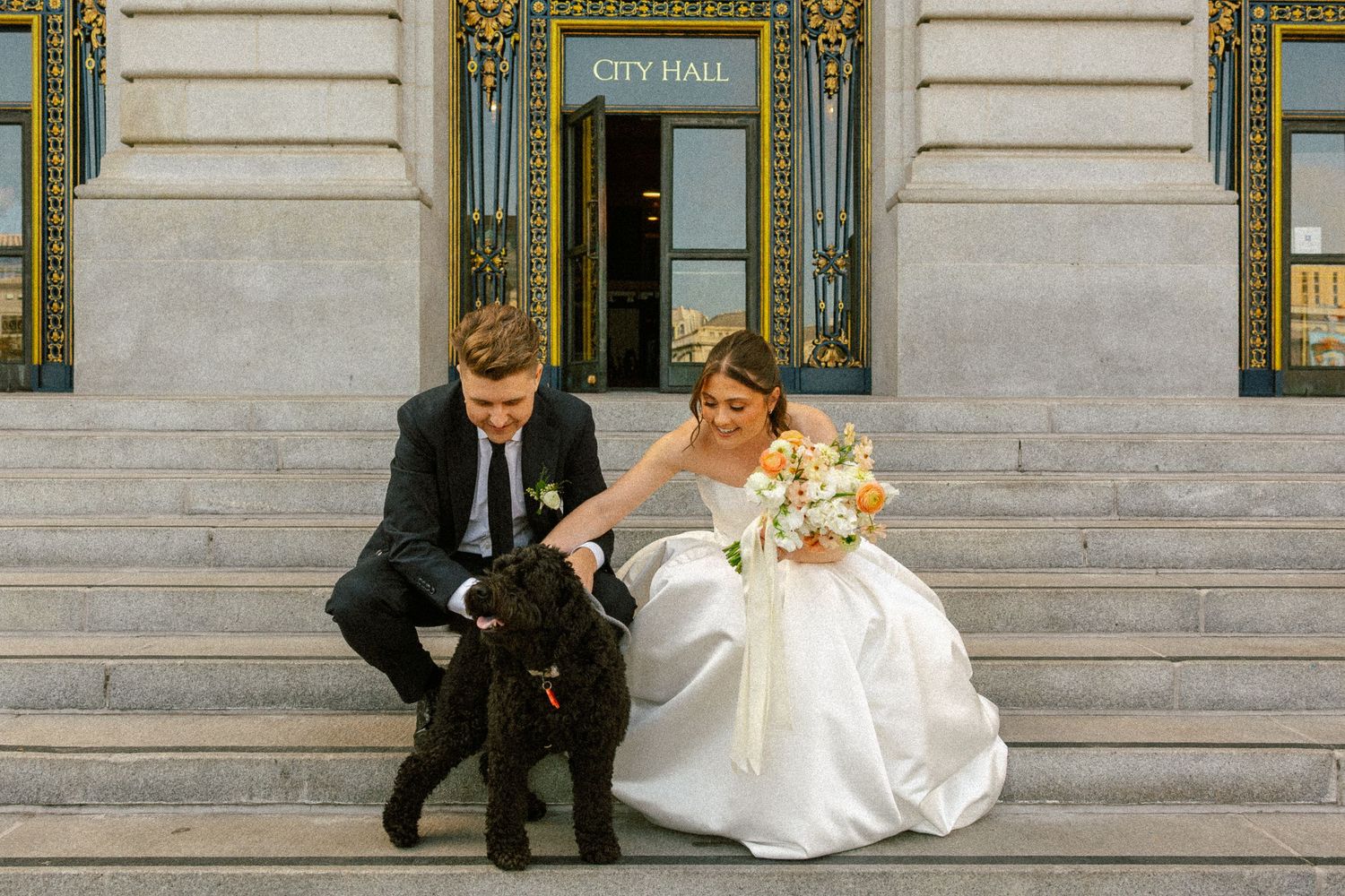 Bride and groom in wedding attire petting a black dog on City Hall steps in San Francisco.