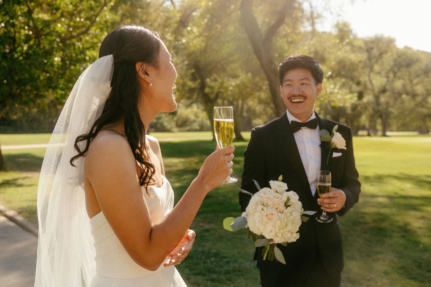 Bride and groom laughing and toasting with champagne glasses outdoors on their wedding day.