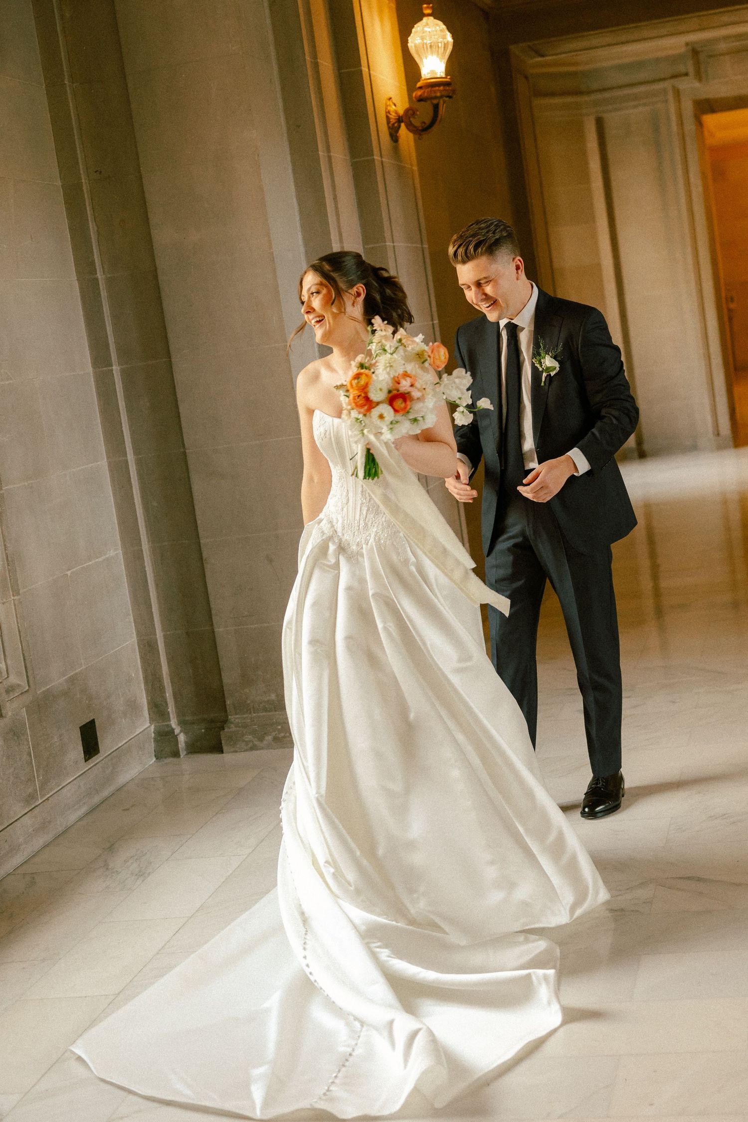 Bride in white gown holds colorful bouquet walking with groom in black suit through elegant marble hallway.