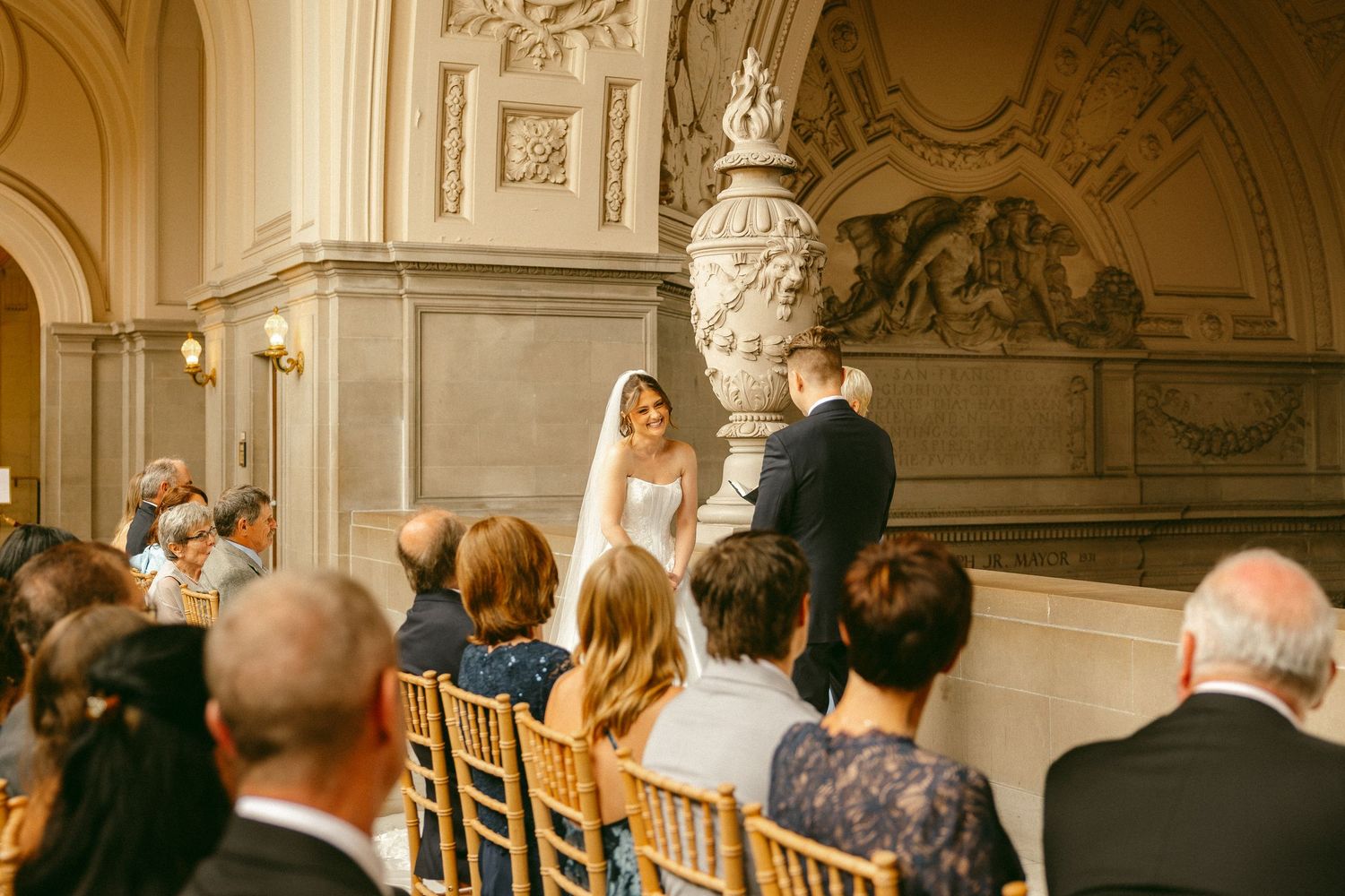 Elegant wedding ceremony at San Francisco City Hall with bride in white gown and veil exchanging vows before seated guests.