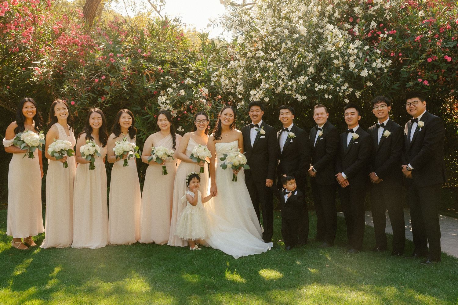 Wedding party posing outdoors with bridesmaids in blush gowns and groomsmen in black tuxedos among flowering trees.