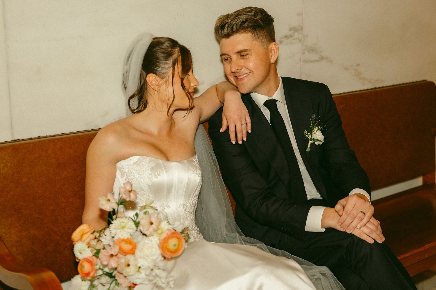 A bride and groom sit together on a vintage bench, sharing a tender moment, with the bride holding a pastel bouquet.