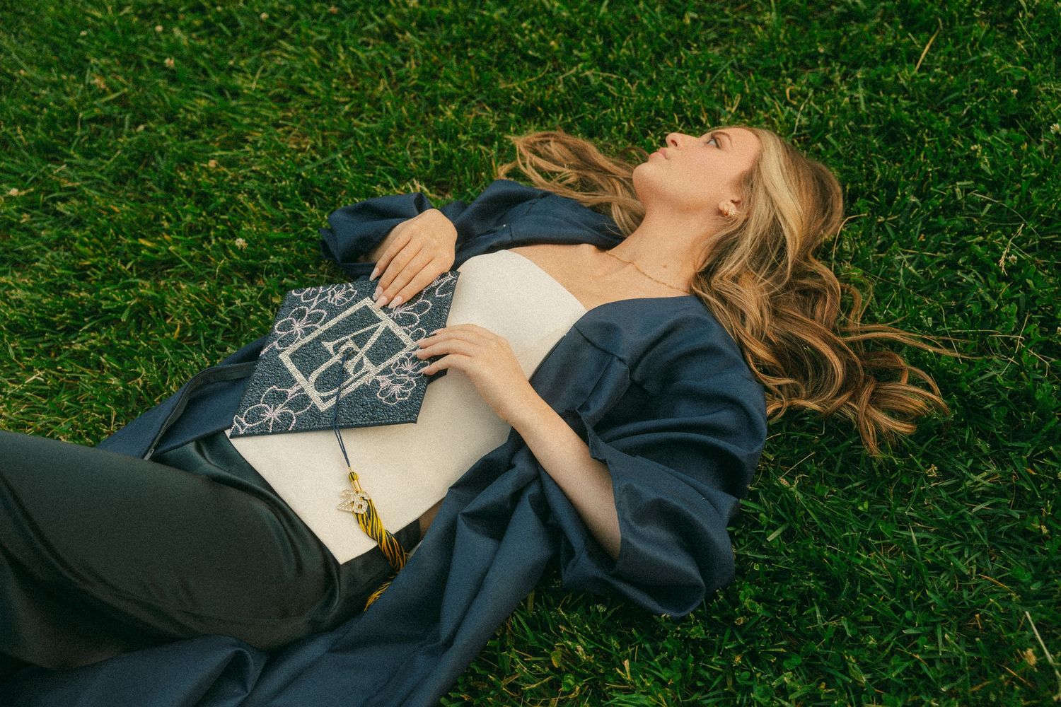 Woman in navy blazer lying on green grass holding a decorative blue and white handbag with gold chain.