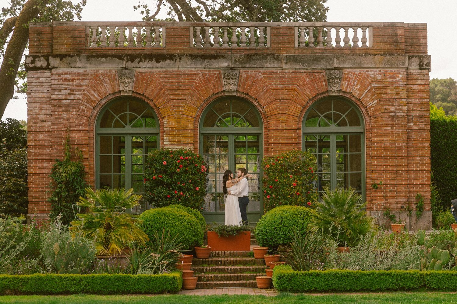 Couple embracing on brick steps before elegant red brick orangery with arched windows and lush garden landscaping.