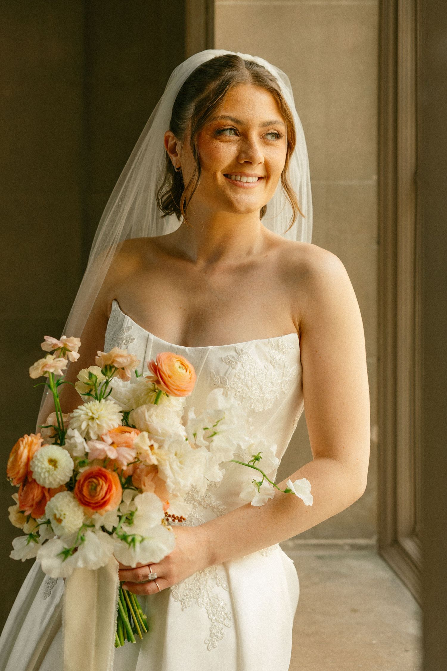 Smiling bride in strapless white gown and veil holds orange and white floral bouquet near a sunlit window.