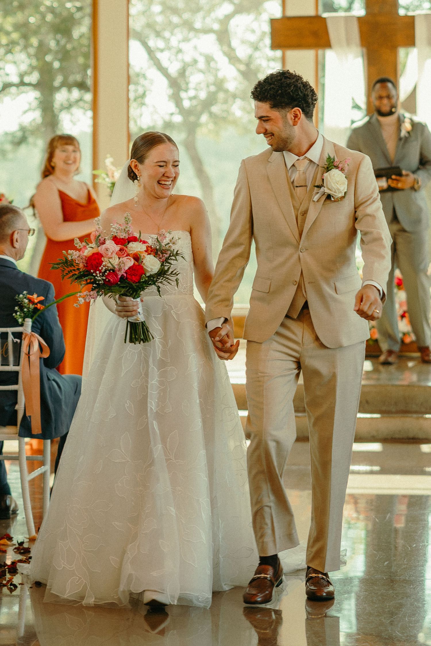 Joyful newlyweds walk down the aisle holding hands, bride in white gown with bouquet, groom in tan suit.
