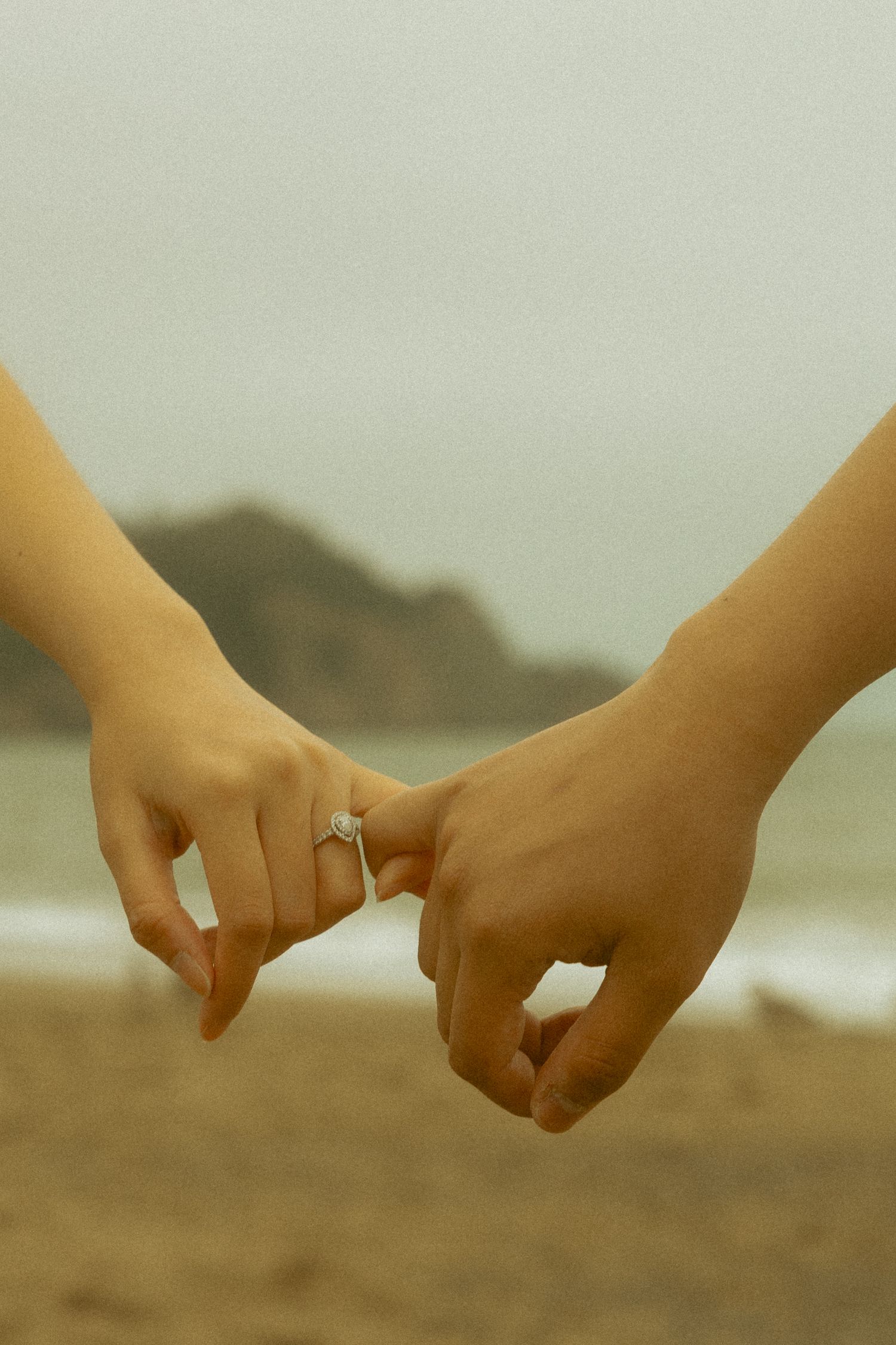 Two hands linking pinky fingers on a beach, one wearing a diamond engagement ring, with ocean waves and cliffs in the background.