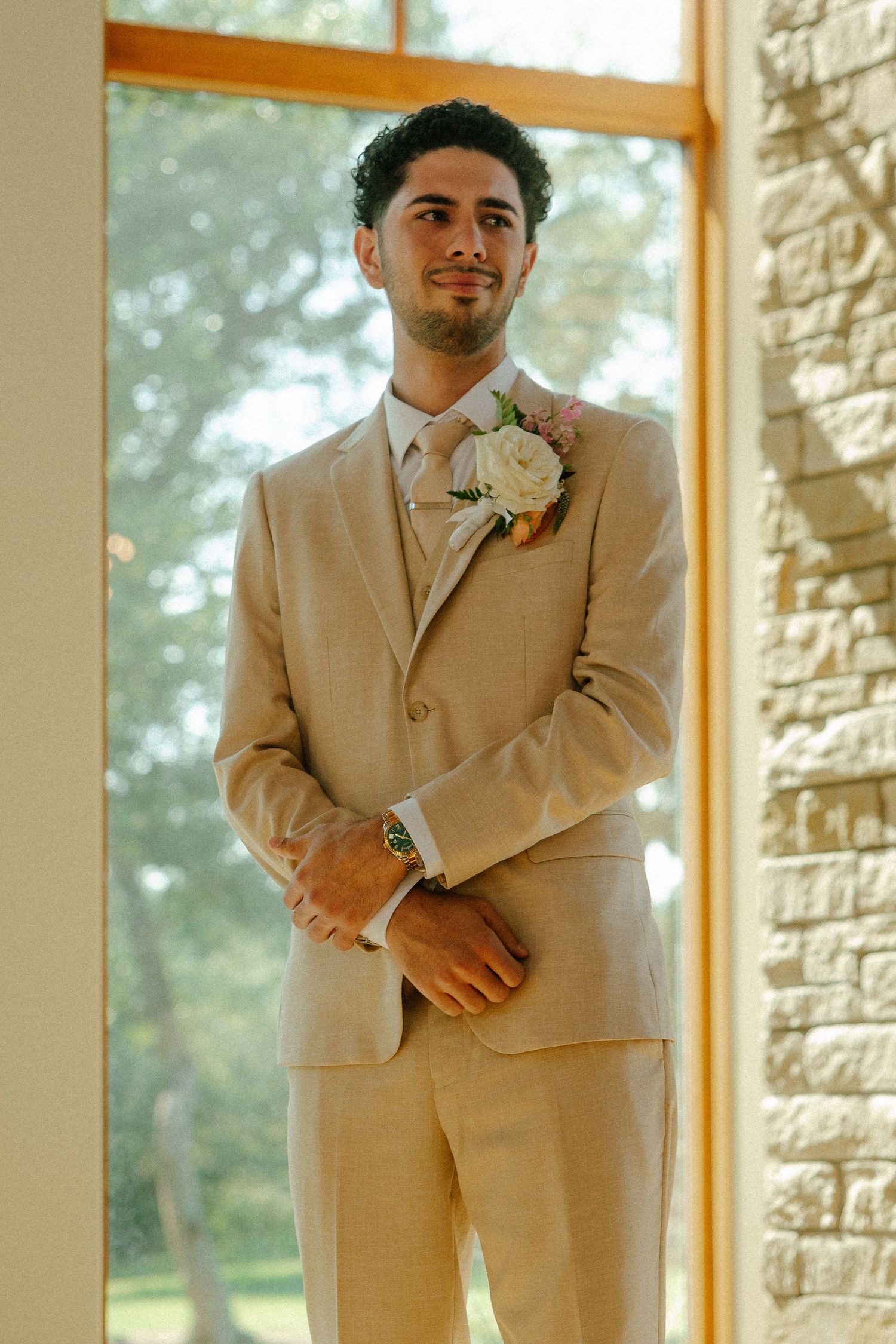 A young groom in a beige linen suit with floral boutonniere stands by a large window at a wedding venue.