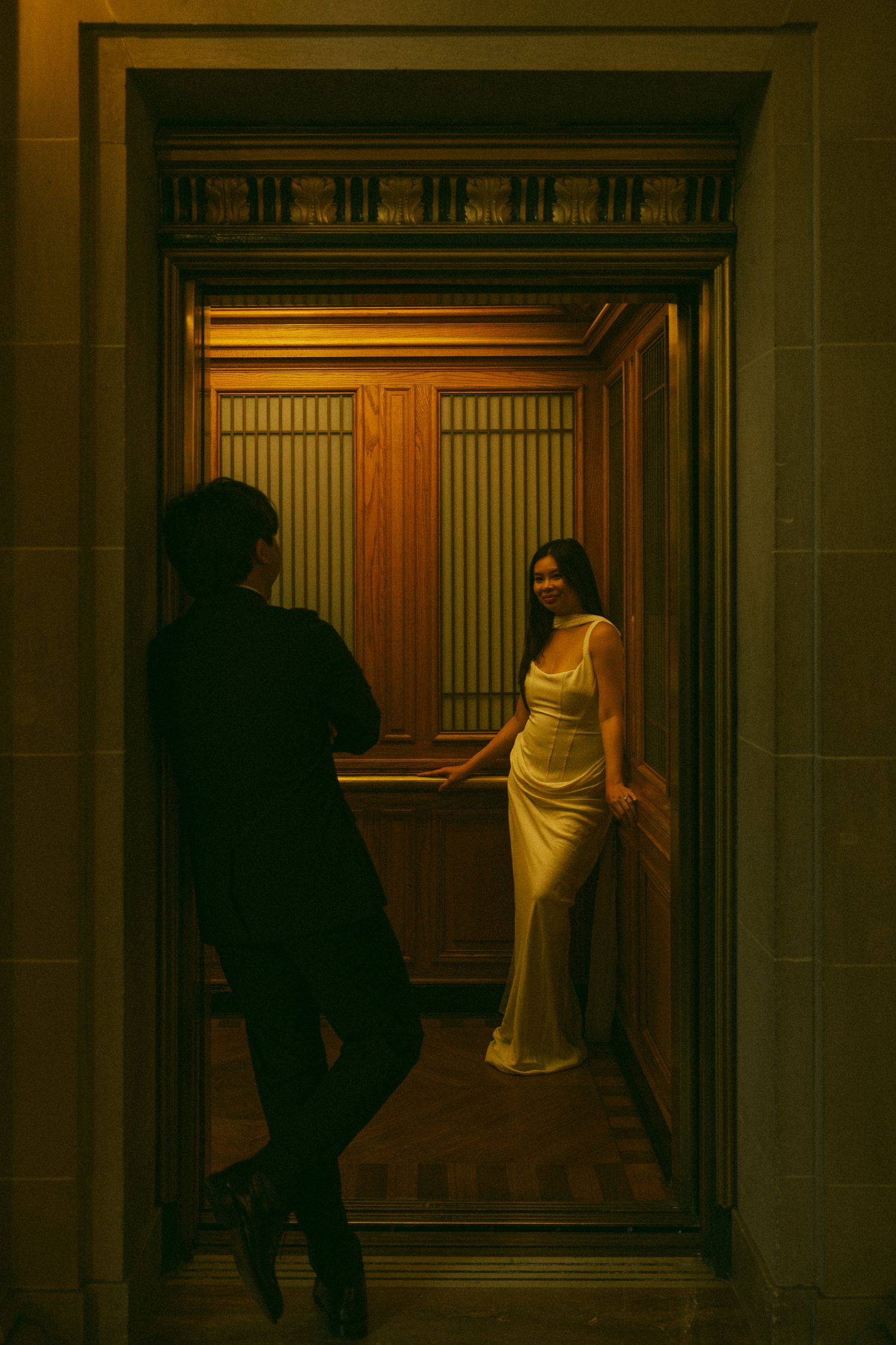 A woman in a gold satin gown poses in an ornate wooden elevator while a man in black photographs her.