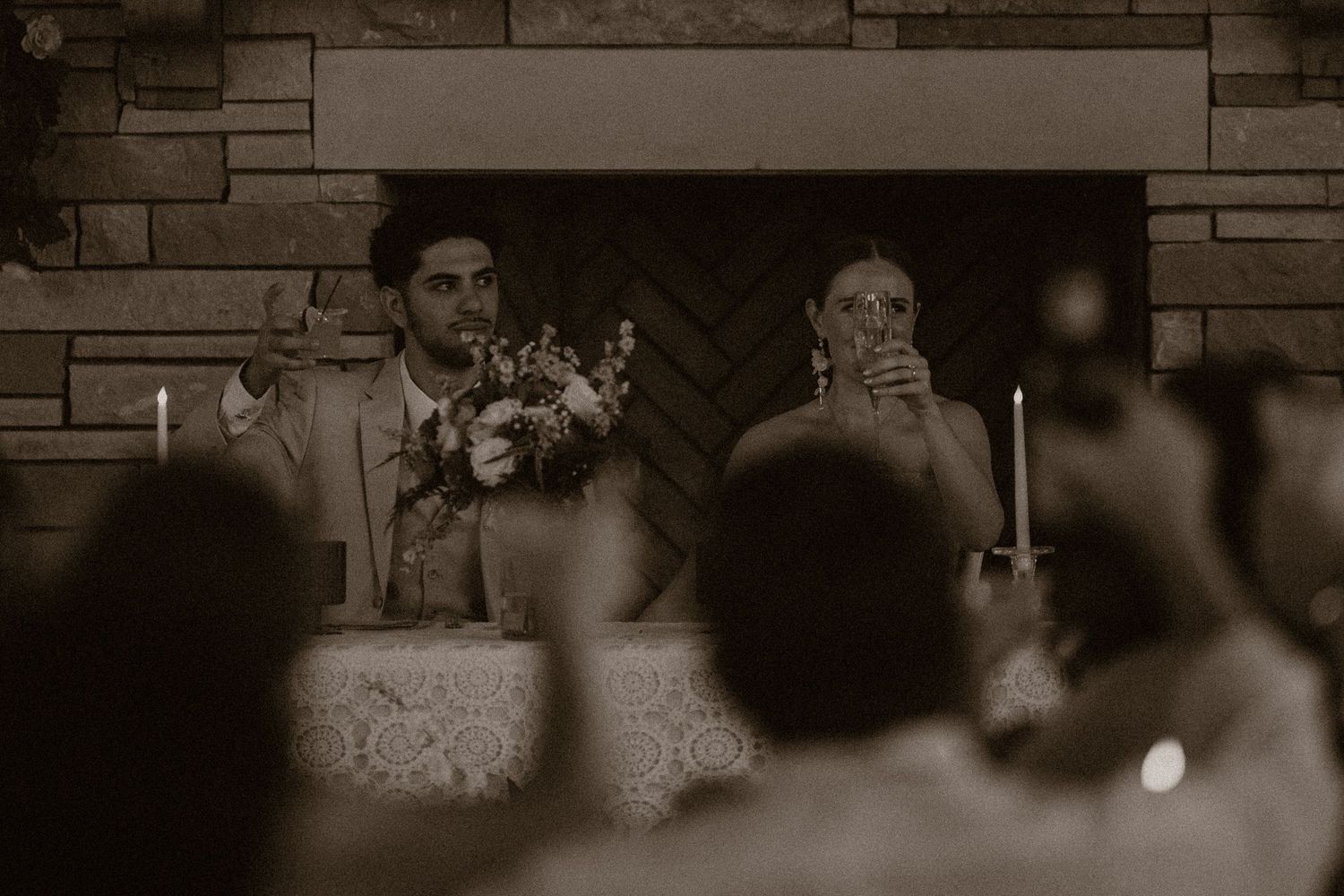 Bride and groom seated at wedding reception table in sepia tone photo with stone fireplace backdrop.