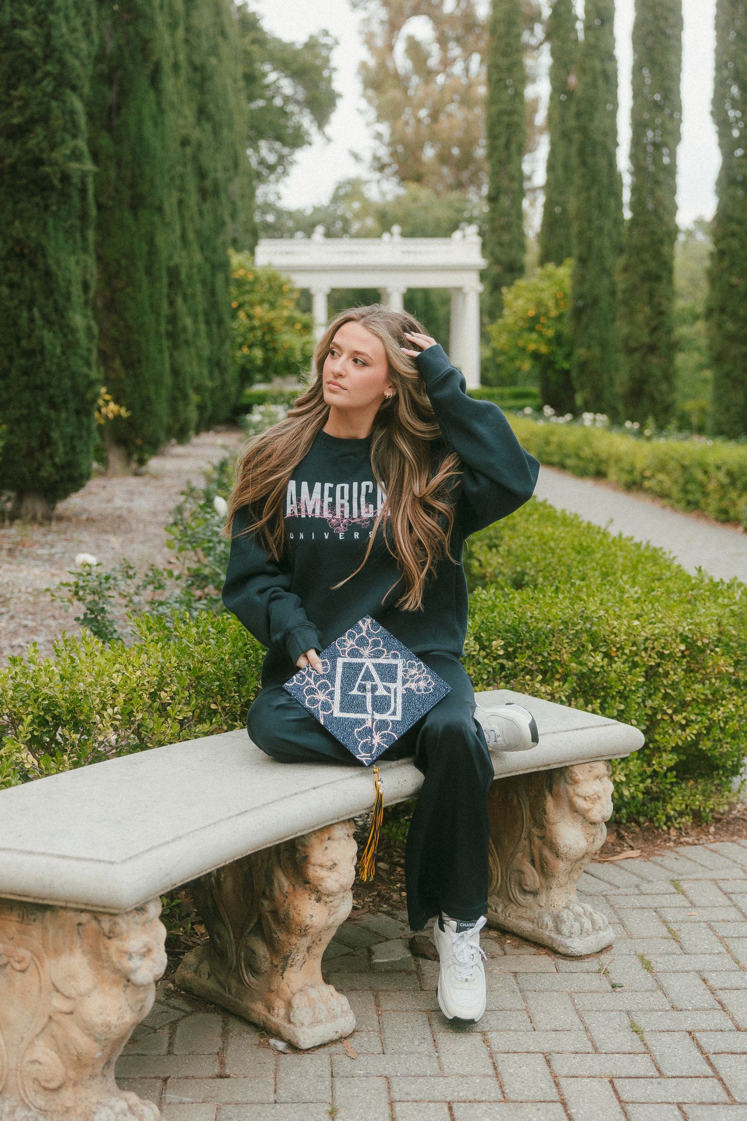 Young woman in American sweatshirt sitting on stone bench holding graduation cap in formal garden.