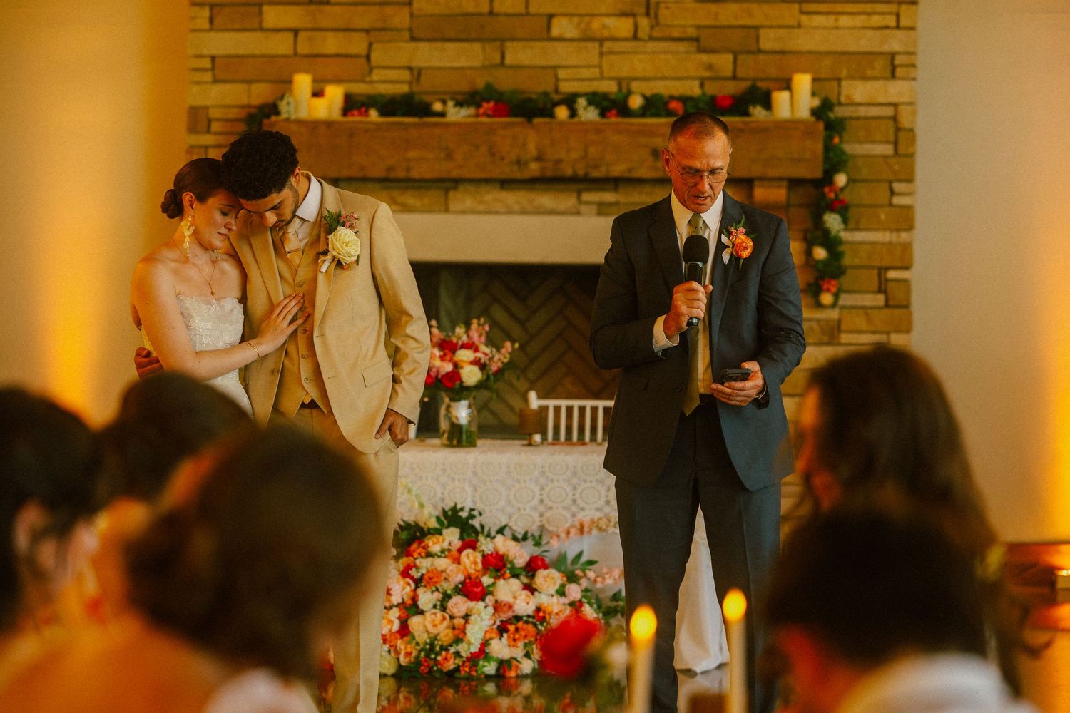 Father of the groom giving a heartfelt wedding speech by a fireplace while the bride and groom listen and smile.
