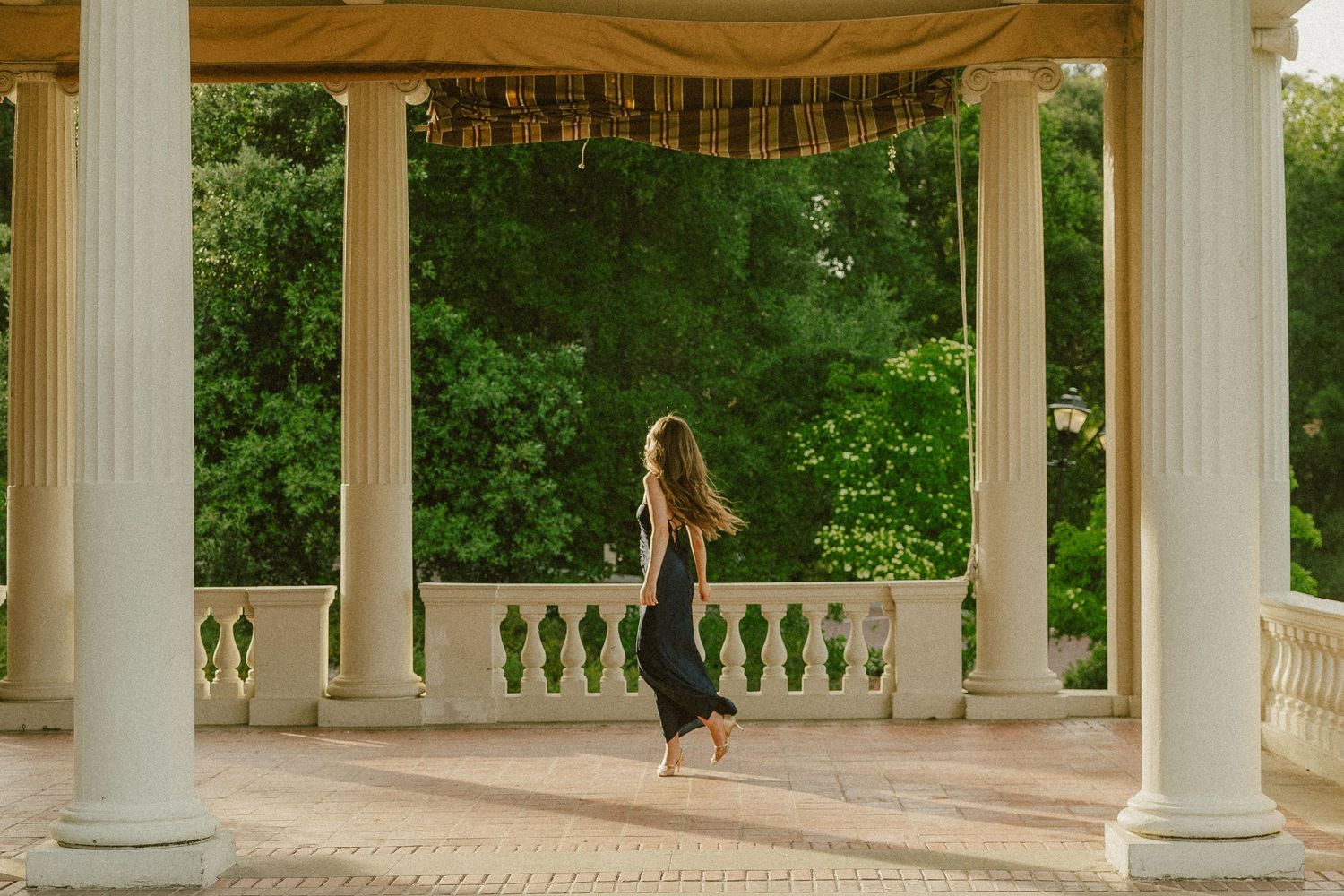 A woman in a black dress walks barefoot through an elegant columned pavilion surrounded by lush green trees.
