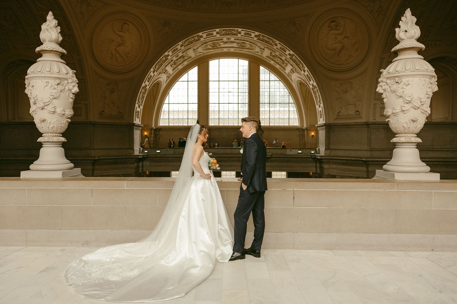 Bride and groom share romantic moment beneath grand arched window with ornate sculptures at San Francisco City Hall.