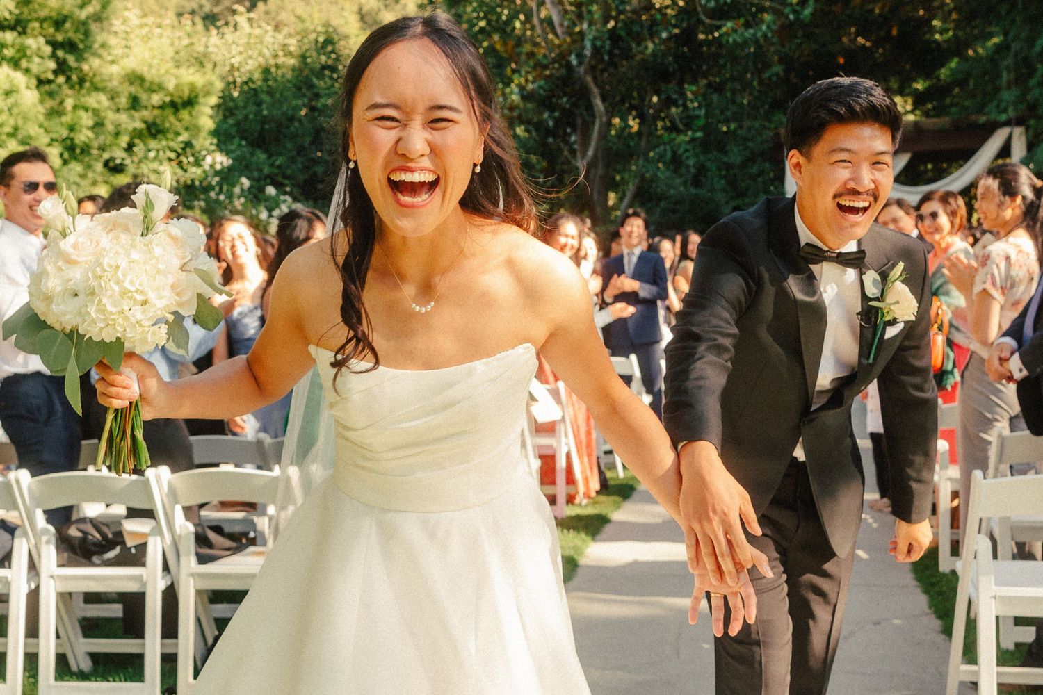 Joyful newlywed couple laughing and holding hands as they walk down the aisle at an outdoor wedding ceremony.