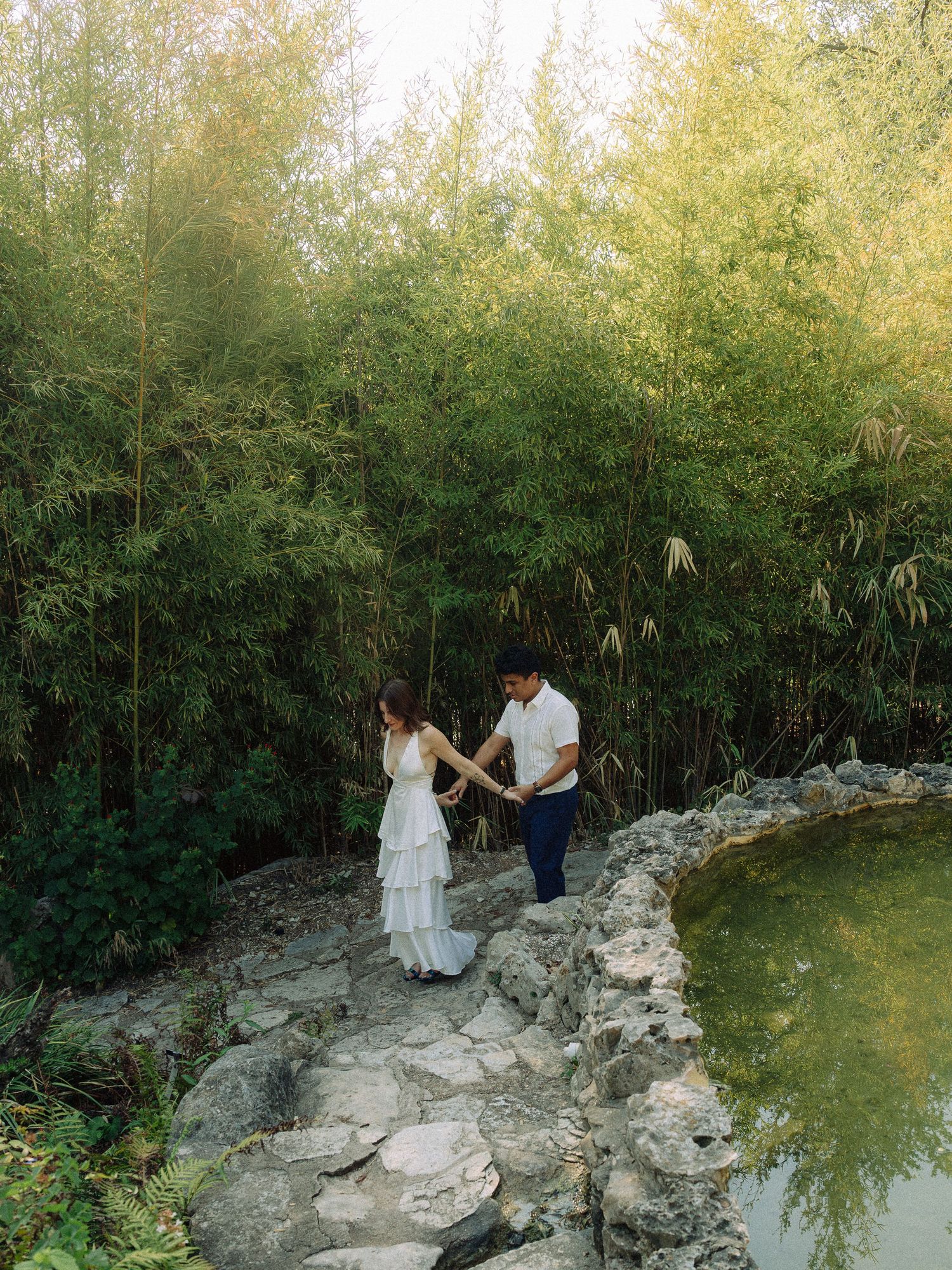 Couple walking along rocky path beside bamboo grove and pond.