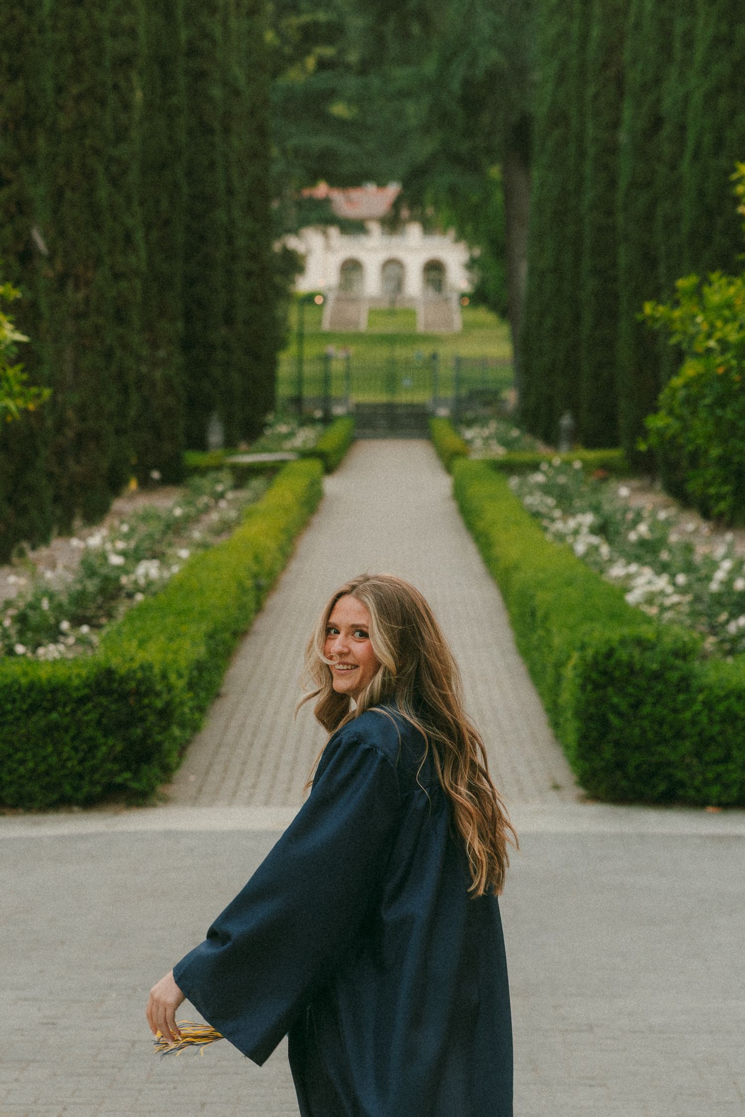 Young woman in navy graduation gown smiles over shoulder in elegant garden pathway with tall cypress trees.