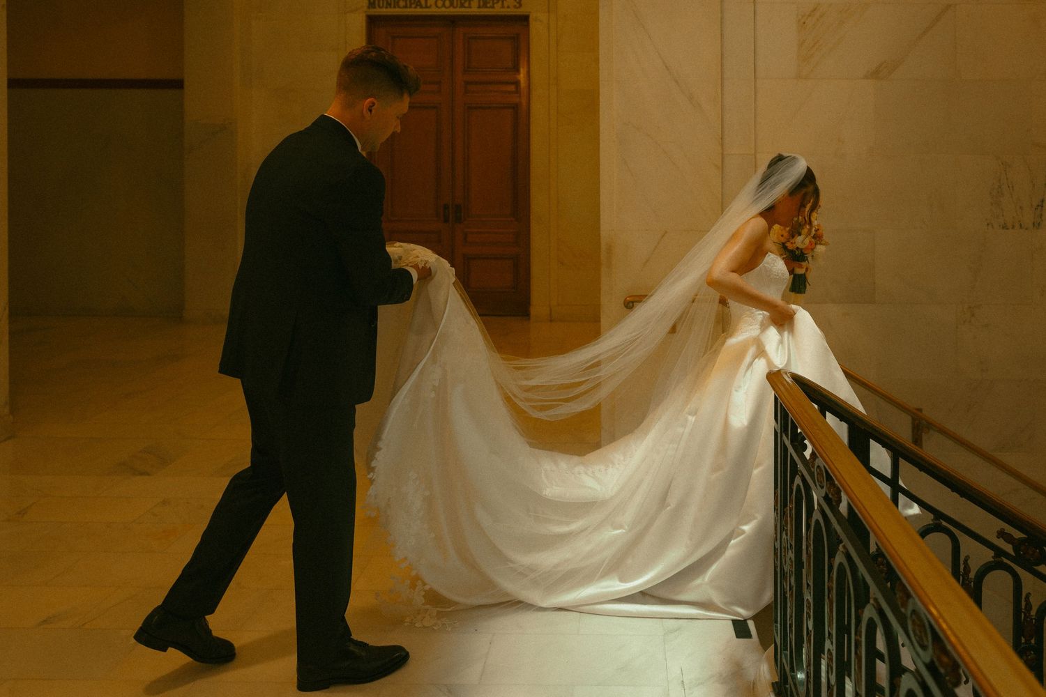 Groom lifting bride's white veil and train on elegant staircase in grand venue during wedding day.