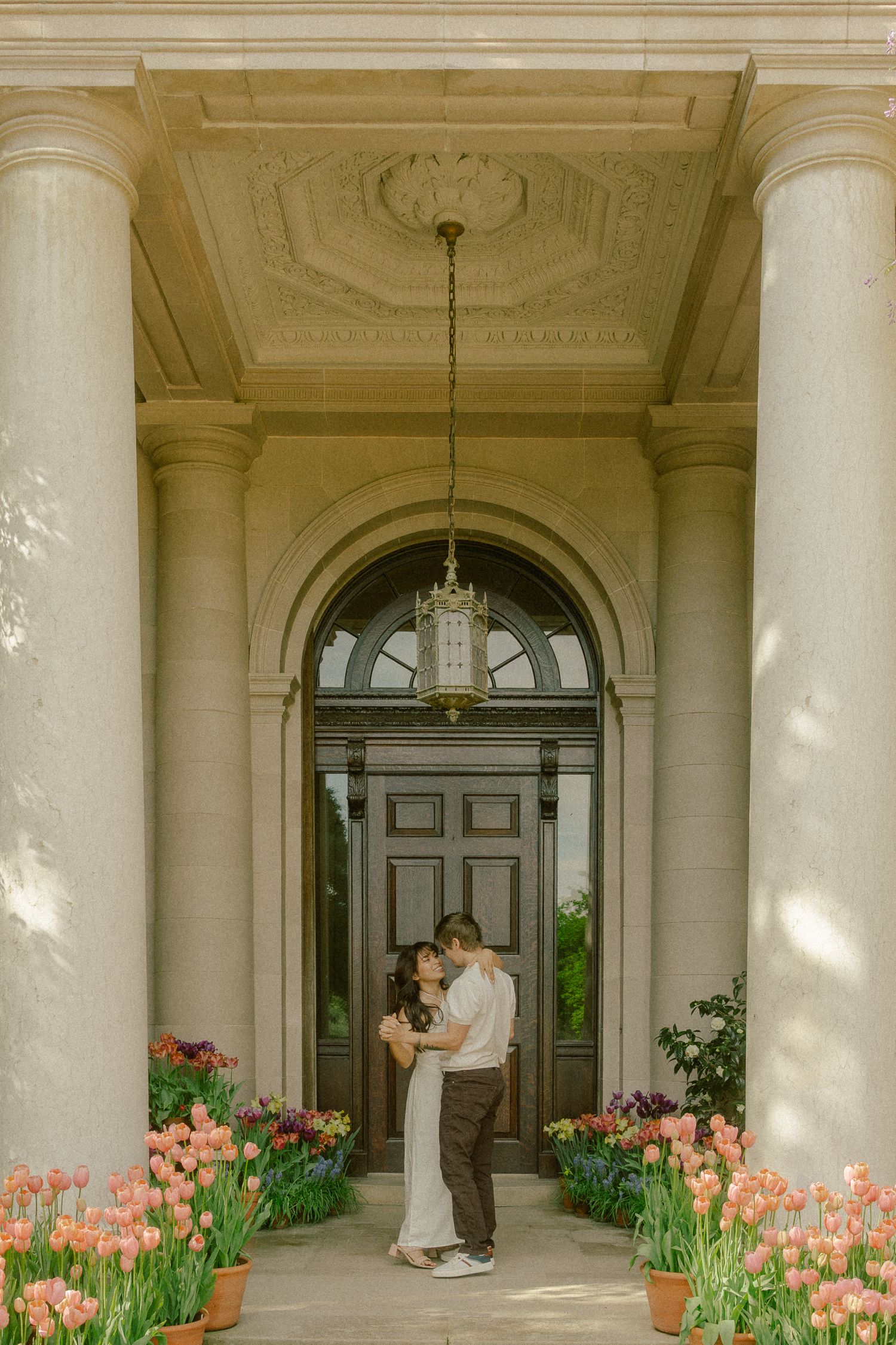 Couple kissing under ornate archway entrance with pink tulips at elegant neoclassical building.