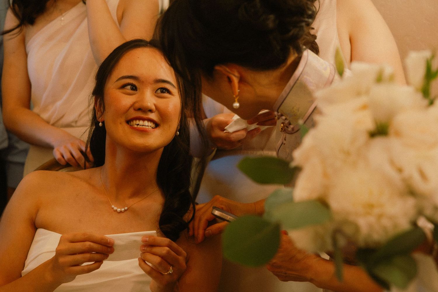 Bride smiling warmly as someone whispers in her ear, holding white flowers at a wedding celebration.
