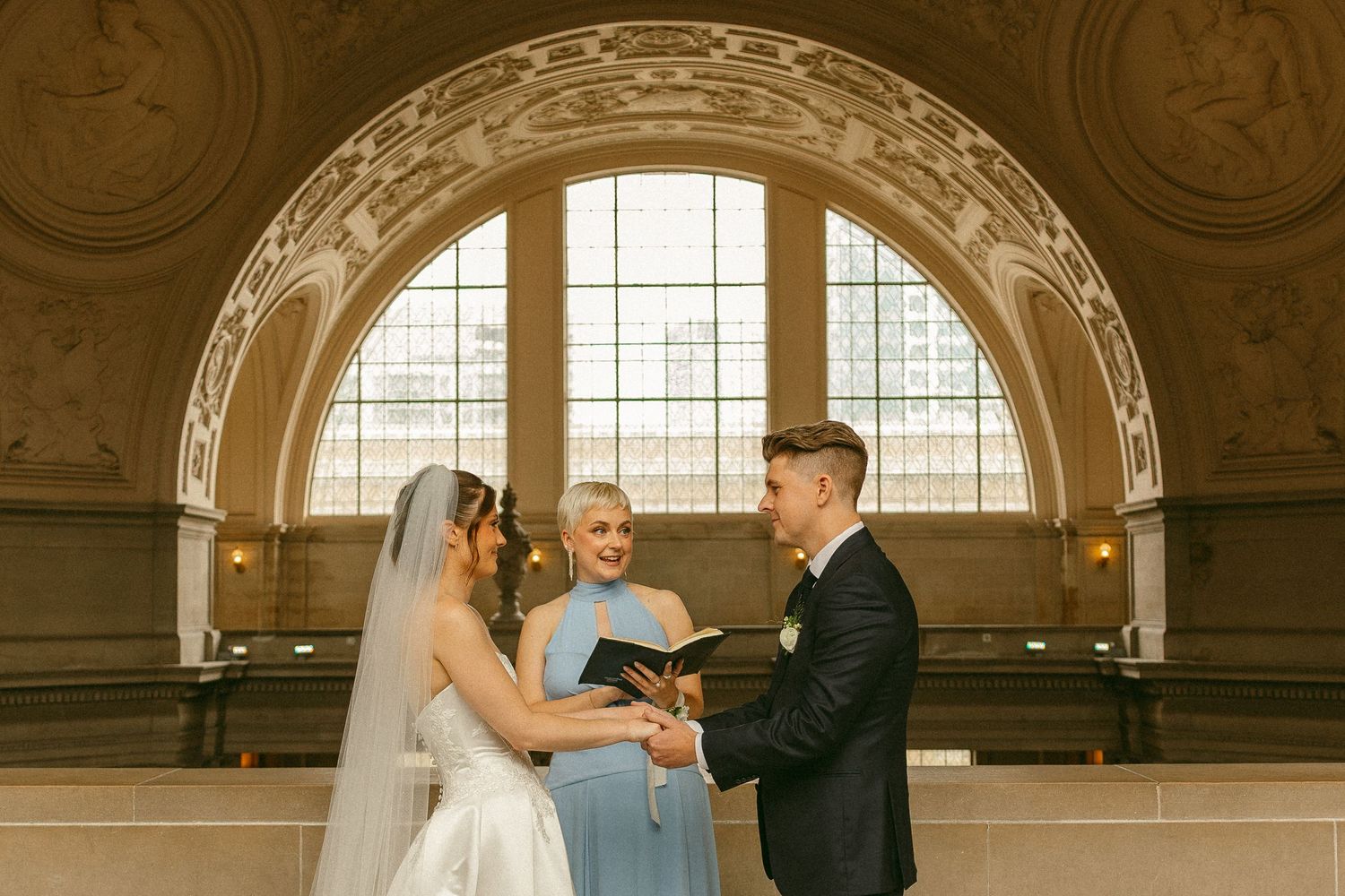 Couple exchanging vows at San Francisco City Hall wedding ceremony with officiant under ornate arch.