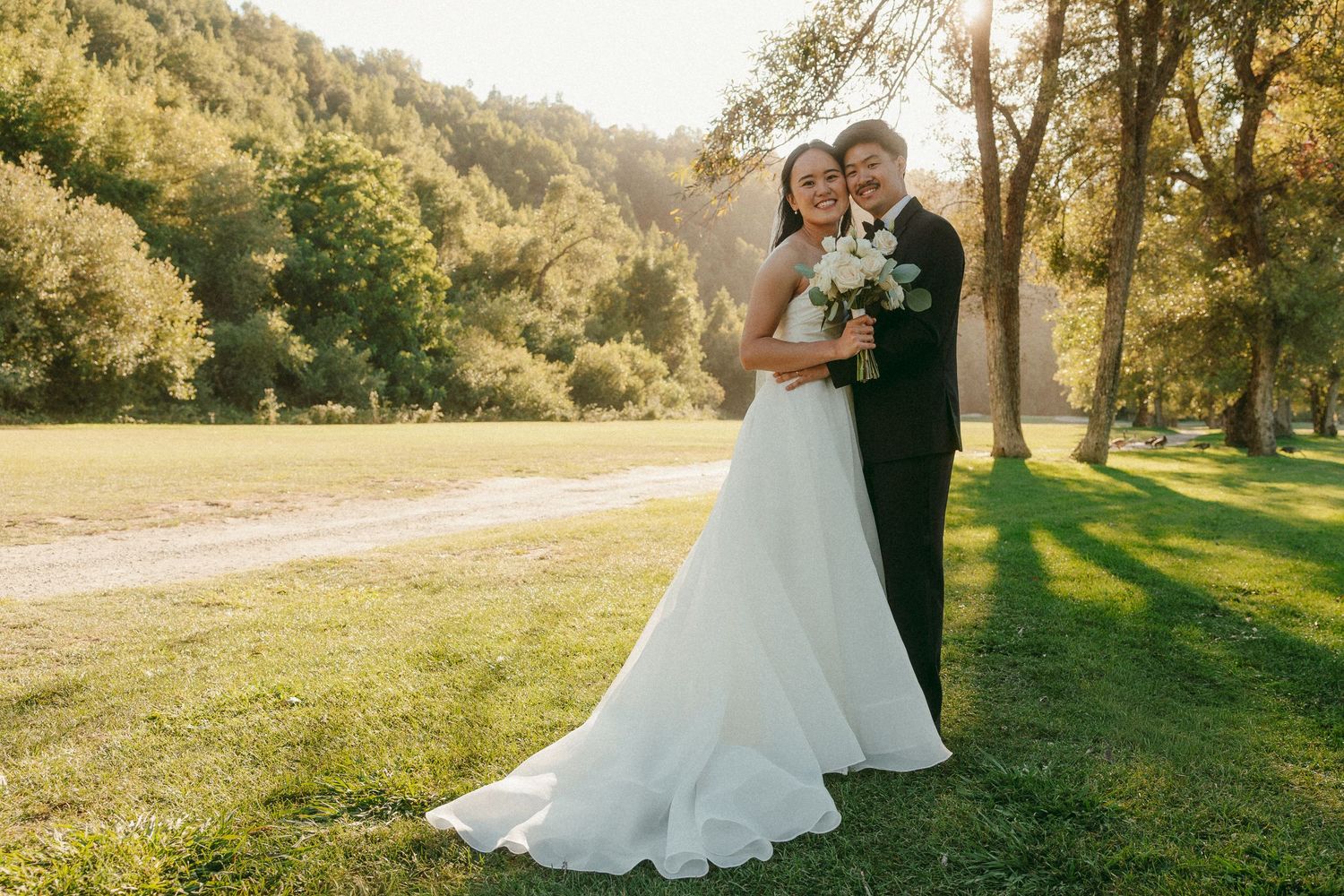 Bride and groom embracing in a sunlit green meadow surrounded by trees, bride holding white bouquet in wedding dress.