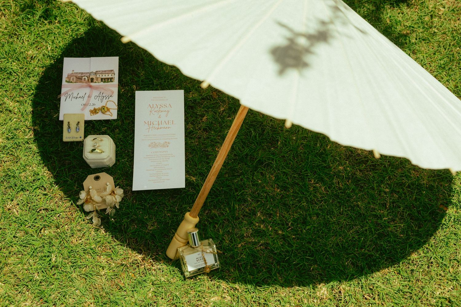 White parasol casting shadow on grass beside wedding stationery, number cards, and decorative items at outdoor ceremony.