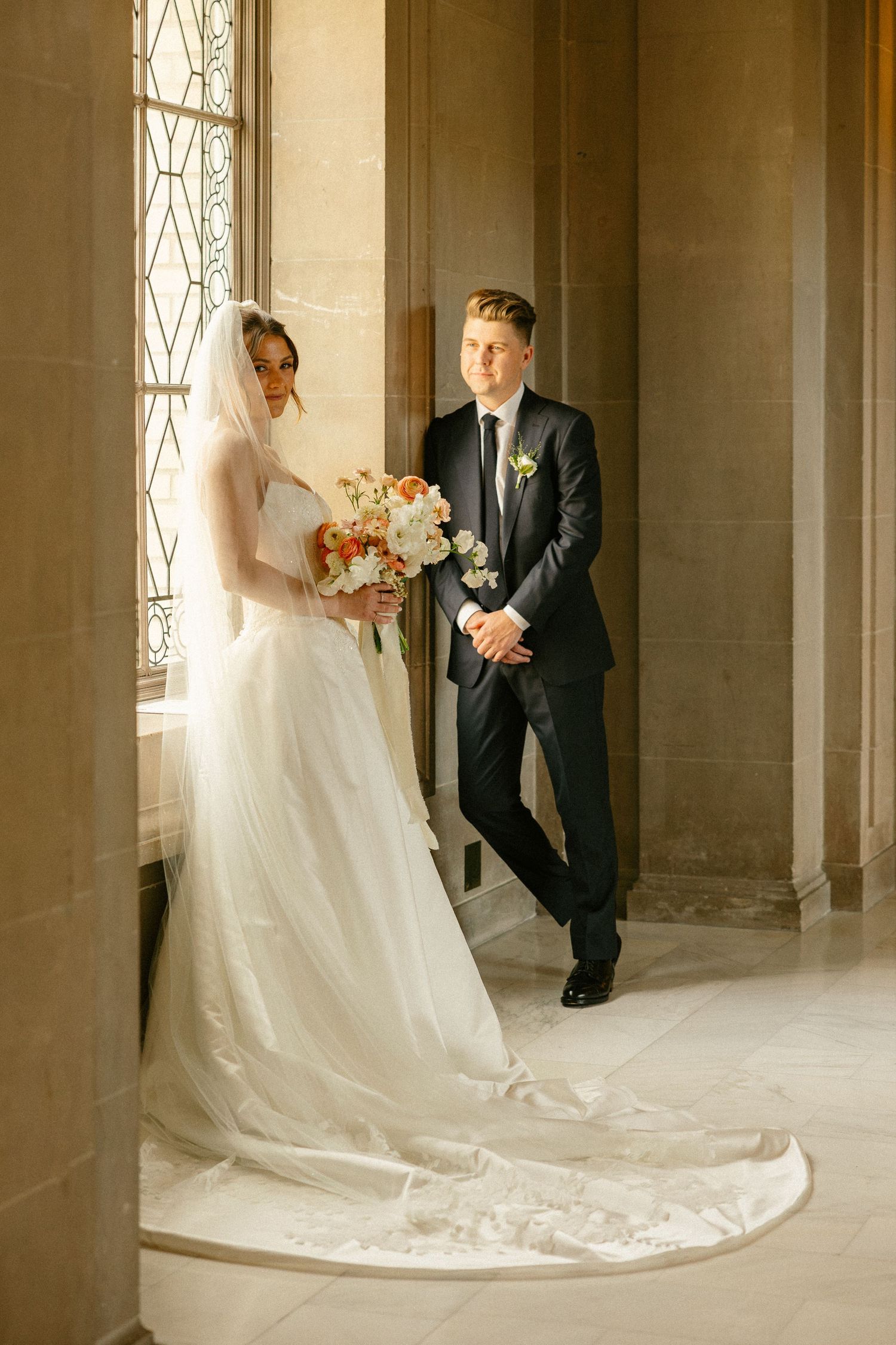 Bride in white gown with veil holding bouquet stands beside groom in black suit near ornate window.