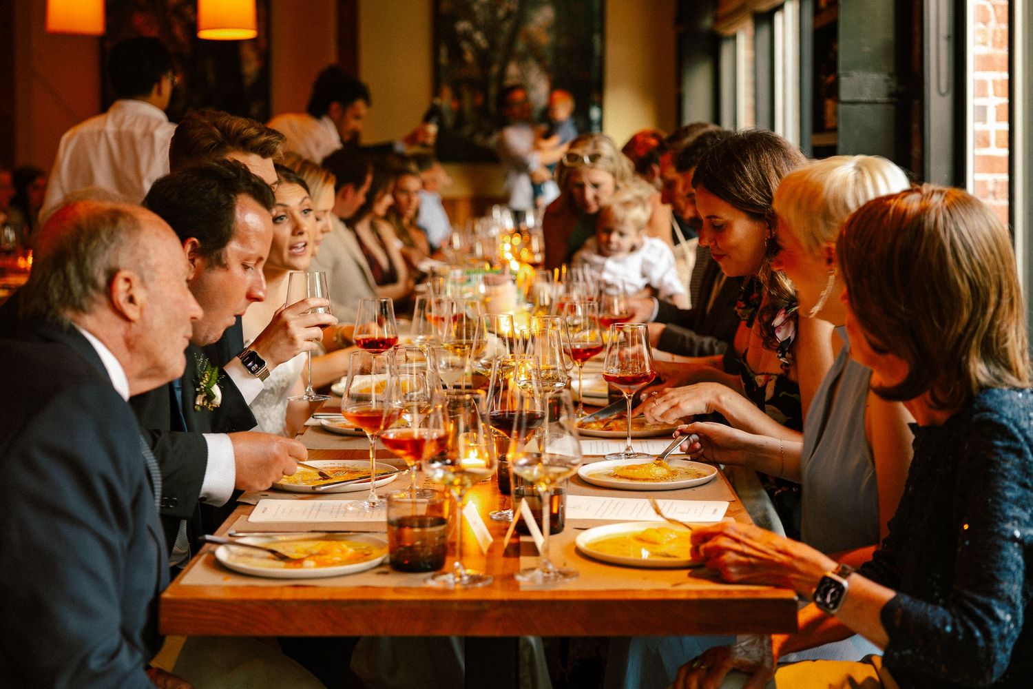 Guests dining at a long candlelit restaurant table with wine glasses, enjoying a formal group dinner in a warm, elegant setting.