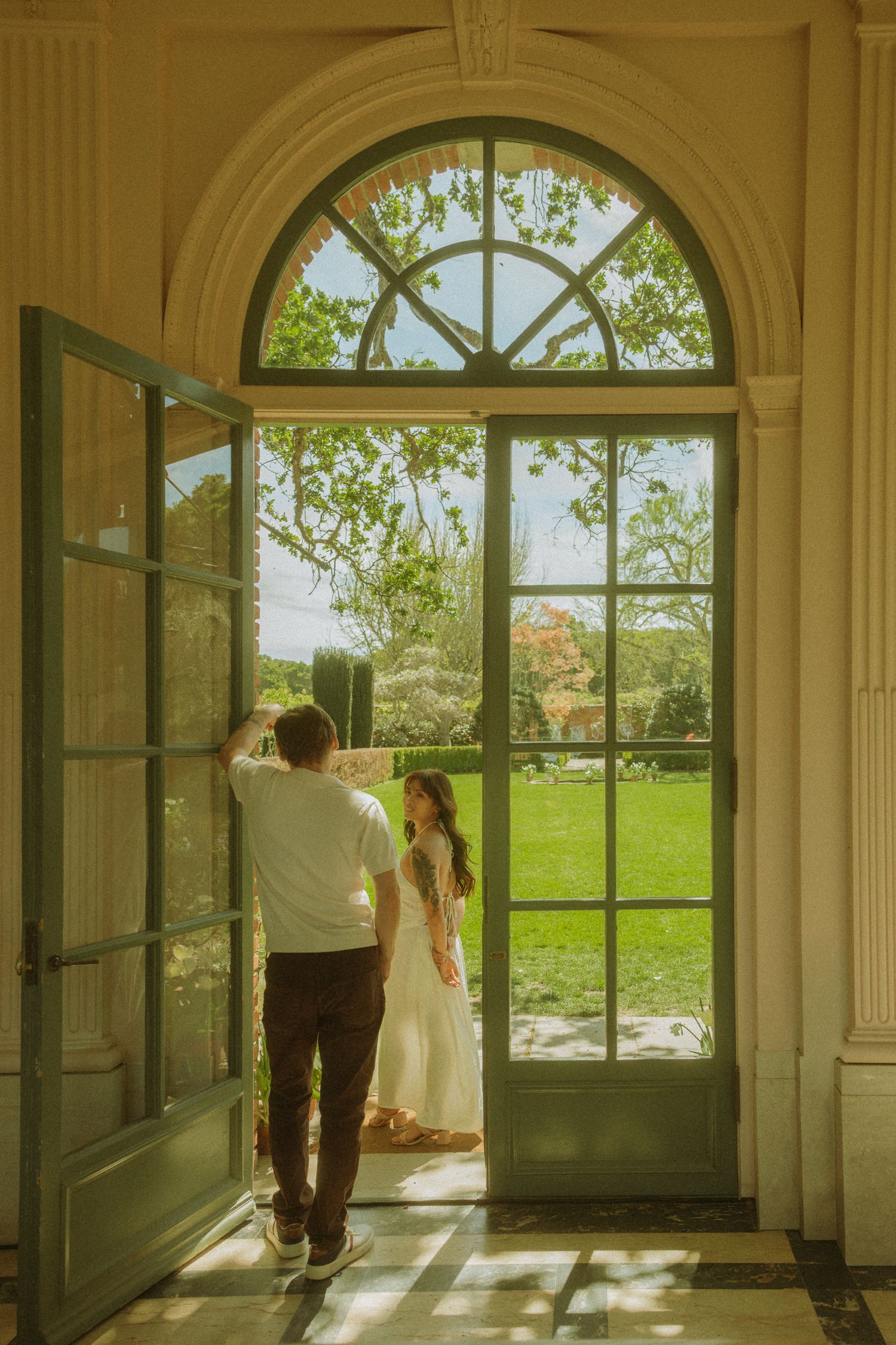 Couple standing in elegant arched doorway overlooking lush green garden, bride in white dress, sunny day.